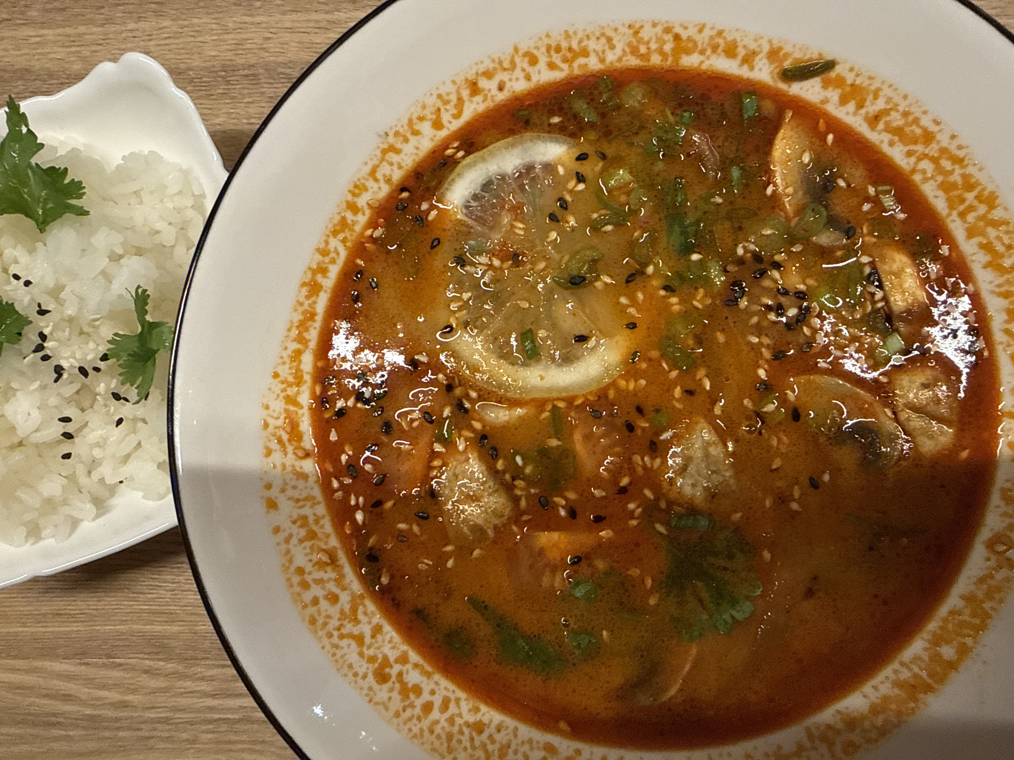 The image displays a meal consisting of a bowl of soup and a side dish of rice, placed on a wooden table.
In the foreground and occupying the majority of the frame, a round white ceramic bowl with a thin black rim contains a reddish-orange broth soup. The soup's surface is garnished with scattered black and white sesame seeds, finely chopped green herbs (likely green onions or cilantro), and two visible thin slices of lemon. Other ingredients partially submerged in the broth include sliced mushrooms and lighter-colored irregular pieces, possibly tofu or chicken. An oily residue, predominantly orange, is visible on the inner rim of the bowl, just above the soup line.
In the upper right portion of the image, partially visible, is a smaller white ceramic dish with an irregular, slightly scalloped edge. This dish holds a serving of white, cooked rice, topped with a sprinkle of black sesame seeds and several small sprigs of fresh green cilantro.
The background consists of a light-brown wooden surface, presenting a vertical grain pattern in the upper left area and a more horizontal grain in the lower right. No individuals are present in the scene. The location is an indoor setting in the city of Unknown, Unknown.