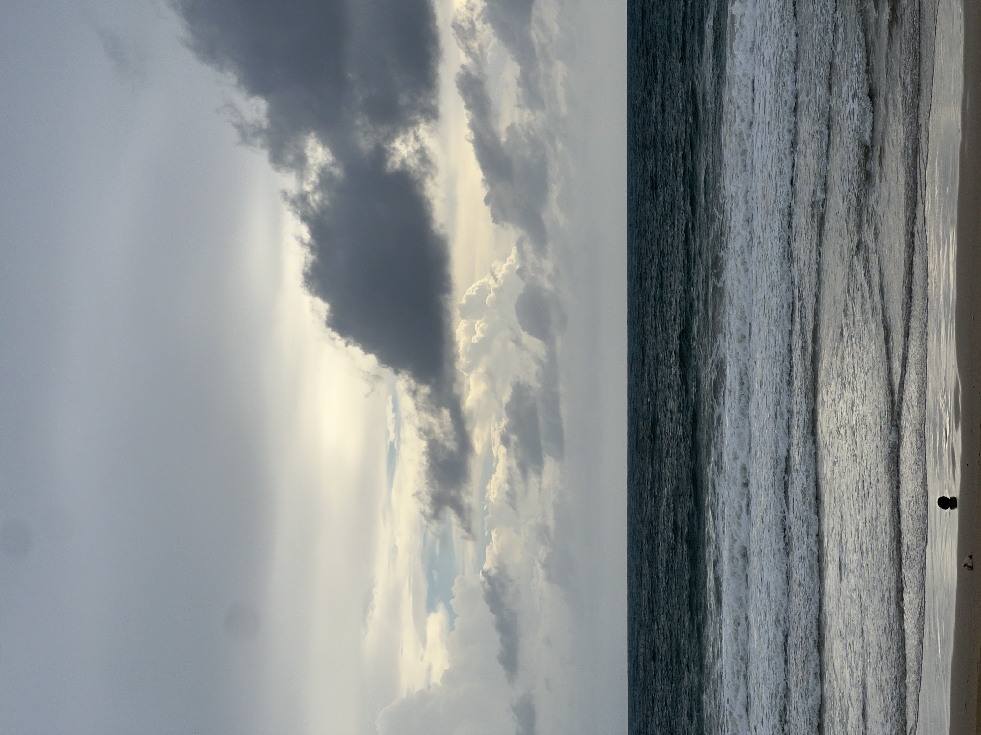 Here is a description of the image:
The image depicts a seascape under an overcast sky. Dark clouds with hints of light breaking through fill the upper portion of the frame, extending down towards the horizon line. The sea occupies the right side of the image, with visible waves creating a textured surface. A small beach is present in the lower right corner, where two dark figures (possibly people) stand near the water's edge.
Location: Unknown, Unknown.
