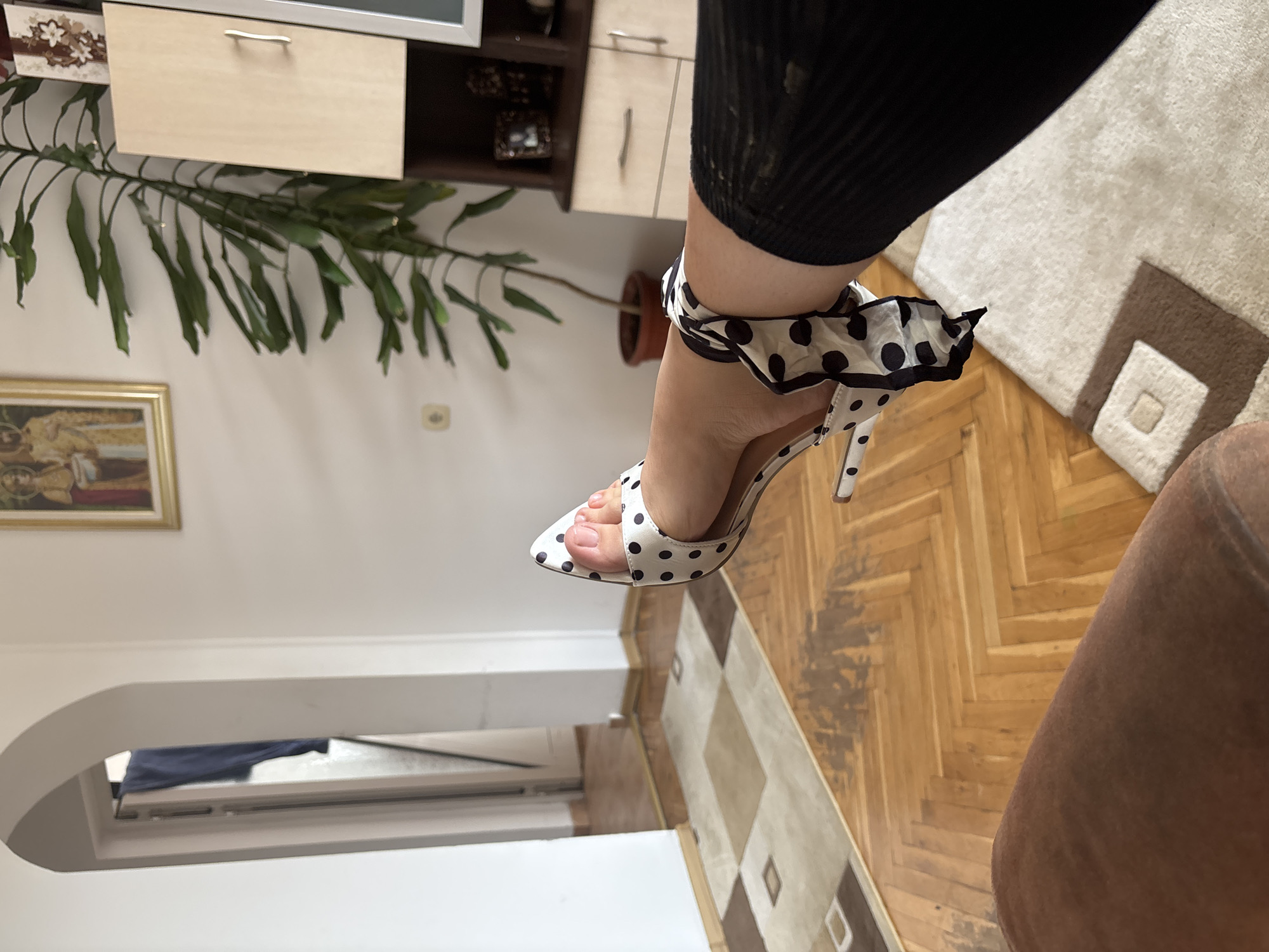 The image shows a close-up of a person's foot wearing a high-heeled shoe with a black and white polka dot pattern. The shoe has a stiletto heel and an ankle strap that is tied with a black and white polka dot scarf. The person is wearing black leggings or stockings. The background is blurry and appears to be an indoor setting with wooden flooring and a rug. There is also a hint of furniture and a plant in the background. The lighting suggests it is daytime. There is no visible text in the image.
