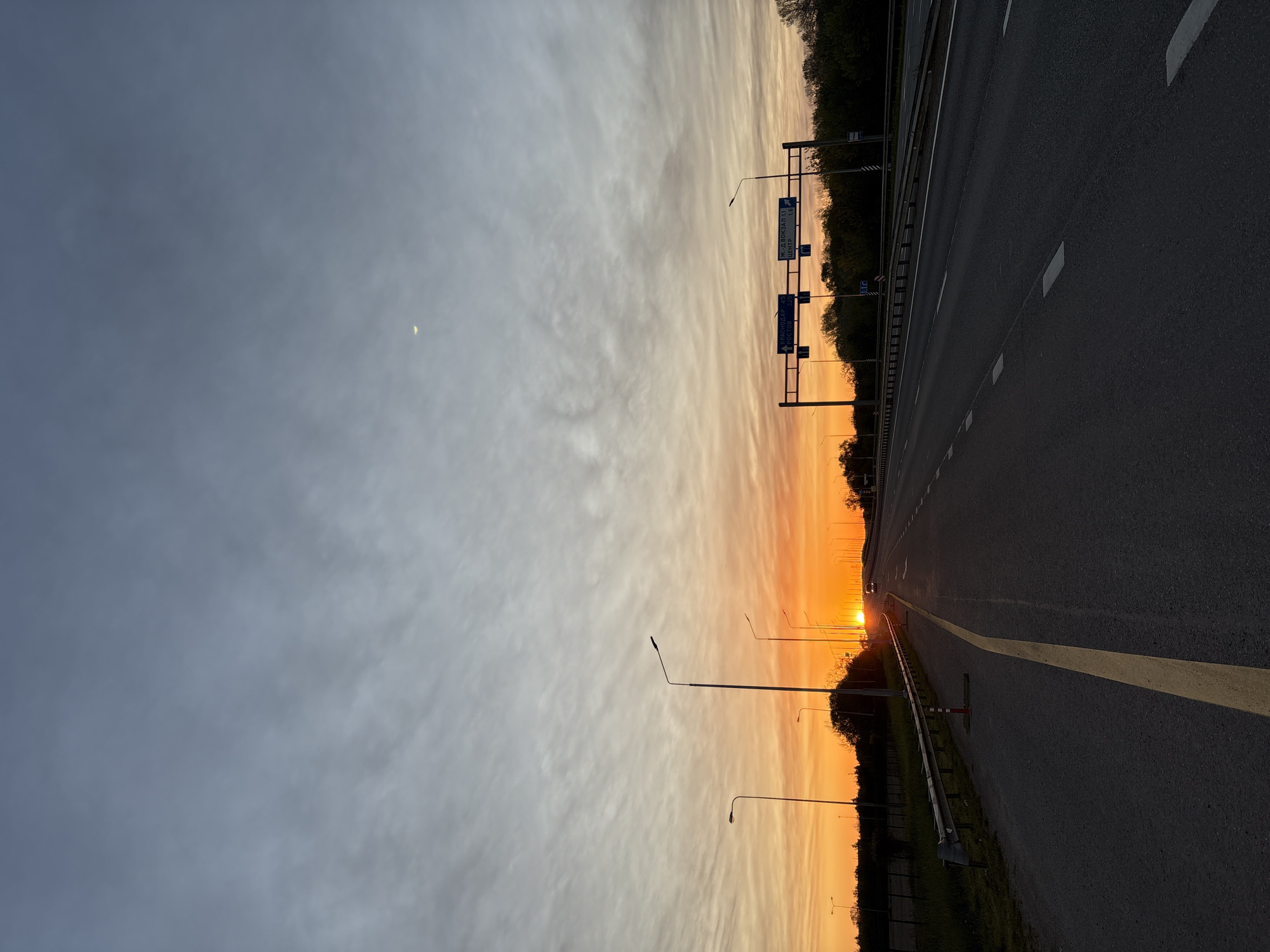 The image depicts a highway scene at sunset in Gelendzhik, Russia. The sky occupies the left side of the frame, transitioning from a dark blue-grey at the top to vibrant orange and yellow hues near the horizon on the right, where the sun is setting. Wispy, horizontally streaked clouds are visible, particularly in the brighter orange sections. A slender crescent moon is present in the upper left, against the darker portion of the sky.
The multi-lane asphalt highway occupies the right side of the image, stretching into the distance. It features white dashed lane markings and a solid yellow line delineating the shoulder. A metal guardrail runs along the far right edge of the road. Several tall streetlights with curved arms are silhouetted against the bright sunset, lining the highway. A dark band of trees or bushes forms the horizon along the right, further emphasizing the golden glow of the setting sun.
A prominent, bright orange sun disk is visible at the horizon, centered towards the right side of the road. Overhead gantries support multiple blue road signs with white Cyrillic text. The top sign indicates directions to "Ж.Д.ВОКЗАЛ 11" (Railway Station, 11 km) and "ЦЕНТР 16" (Center, 16 km). Below it, another sign points towards "КРАСНОДАР 15" (Krasnodar, 15 km) and "РОСТОВ 20" (Rostov, 20 km), confirming the Russian location. The scene conveys the peaceful atmosphere of an evening drive on an open road.