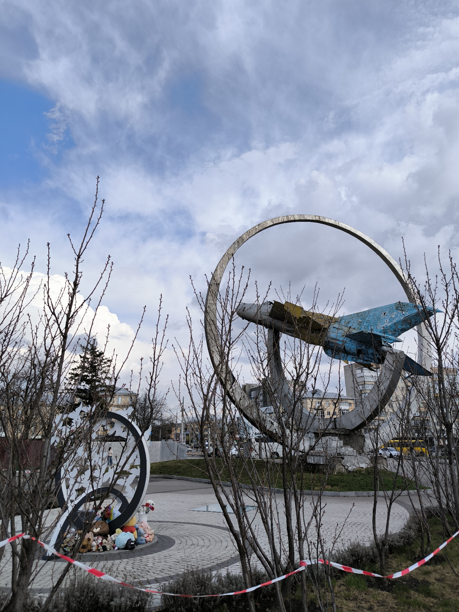The image depicts a memorial scene in Vinnytsia, Ukraine. The main subject is a decommissioned Soviet-era fighter jet mounted within a large, circular metal frame. The aircraft shows signs of age and deterioration, with faded paint and patches. In the foreground, obscured by bare tree branches, is another memorial structure decorated with toys and flowers, encircled by red and white barrier tape. The ground is paved with stone tiles. Buildings are visible in the background, indicating an urban environment. The sky is overcast with varying cloud cover. The memorial appears to be a place of remembrance, likely associated with the ongoing conflict or past tragedies.