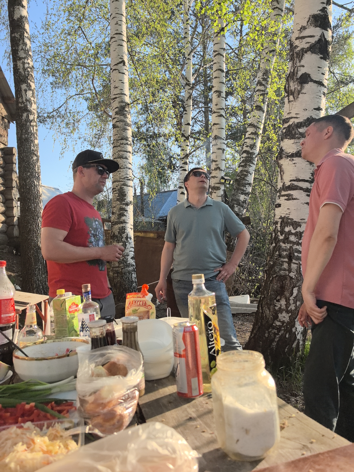The image depicts three men standing around a table laden with food and beverages in an outdoor setting. The location is identified as Noviy, Russia. The men appear to be engaged in conversation. One man is wearing a red t-shirt, a black cap, and sunglasses. Another man is wearing a green polo shirt and sunglasses. The third man is wearing a pink shirt. The table is covered with various food items, including salads, vegetables, and what appears to be bread in a plastic bag. Several bottles and jars of condiments and beverages are also visible. The background shows birch trees and a partial view of a building constructed of logs. The scene is illuminated by sunlight.