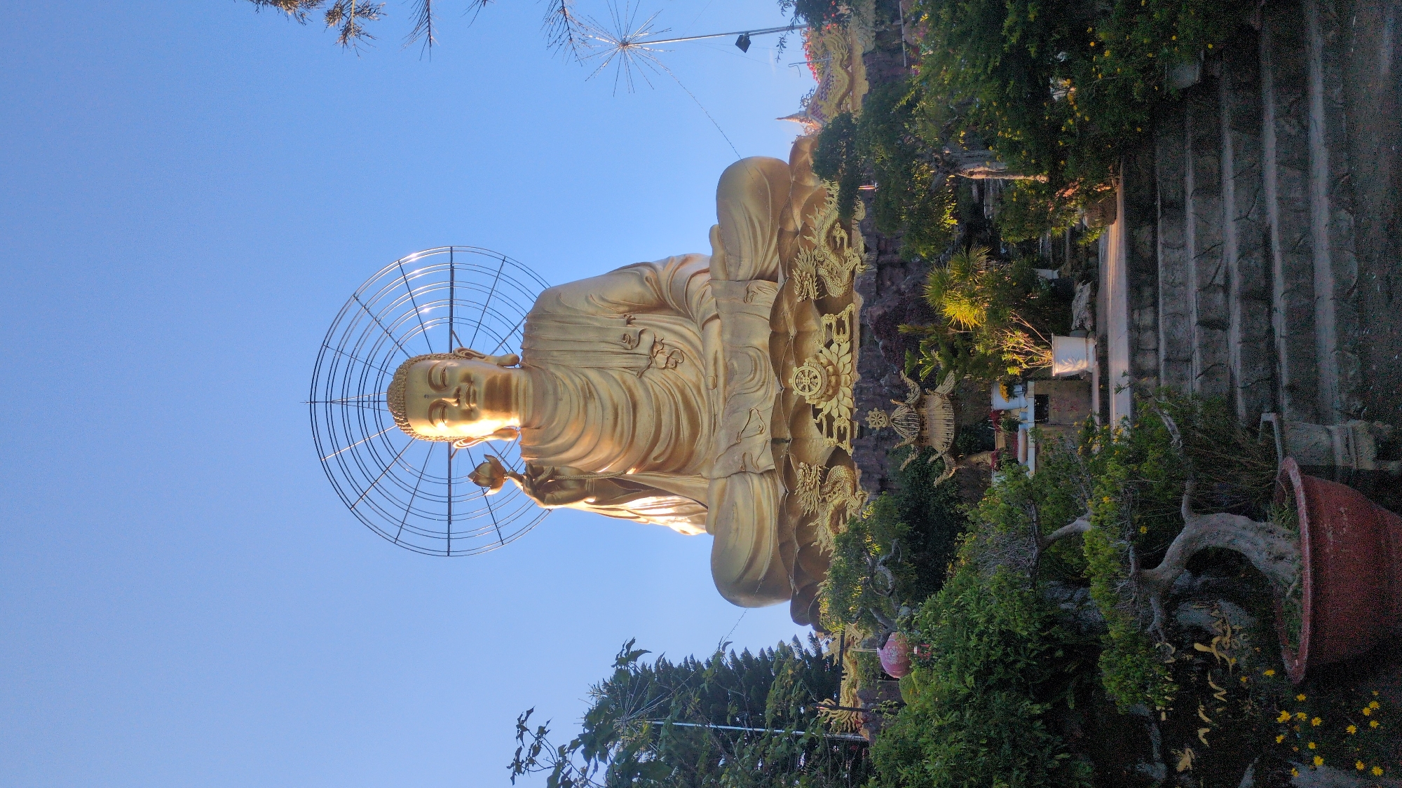 A large, golden Buddha statue sits serenely on a tiered platform, its right hand holding a lotus flower. The statue is adorned with a halo of metallic rings behind its head. The platform itself is decorated with intricate carvings, including dragons and Buddhist symbols. The statue is situated outdoors on a hillside, surrounded by lush green trees, potted plants, and stone steps leading upwards. In the background, the clear blue sky is visible. The scene appears to be a religious or cultural site, possibly a temple complex, located in Lâm Viên - Đà Lạt, Vietnam, as suggested by the architectural style and the presence of Buddhist imagery.
