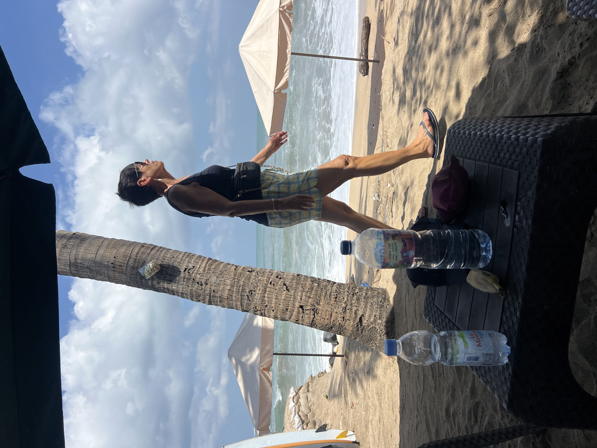 A woman walks along a sandy beach in Kuta, Indonesia, with the ocean and blue sky with clouds in the background. She is wearing a black tank top, plaid shorts, sunglasses, and flip-flops, and has a small black bag slung across her body. In the foreground, on a dark woven table, are two plastic water bottles, a banana, and a maroon hat. A large palm tree is to the left of the woman, and two beach umbrellas are visible in the mid-ground. Waves are breaking on the shore, and sandbags are piled up near one of the umbrellas.
