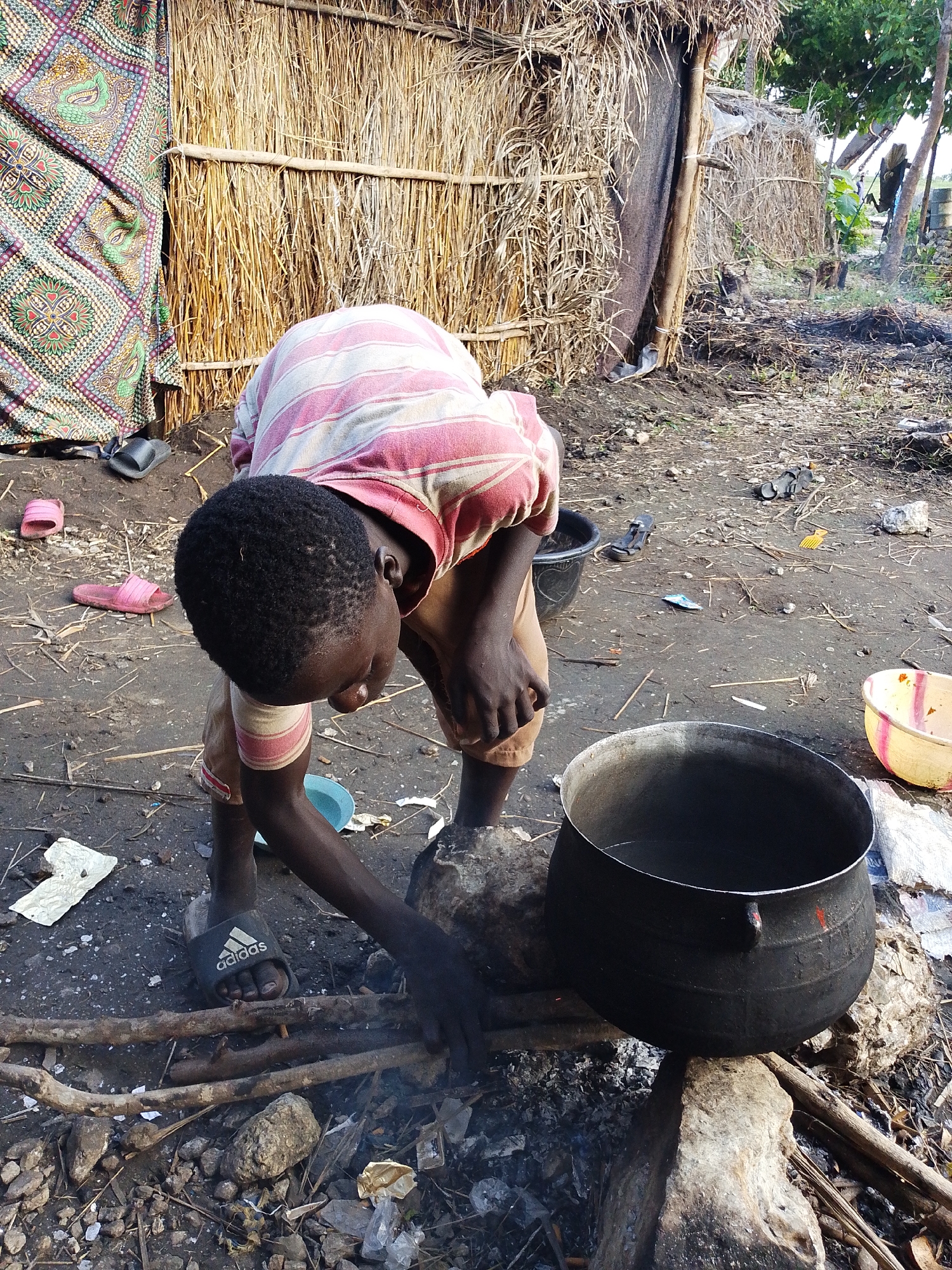 In Kajola, Nigeria, a young boy kneels beside a large black pot on a makeshift stove of rocks and branches, tending to a fire beneath it. He wears a pink and white striped shirt and brown shorts, with one black Adidas sandal on his left foot. The scene is set in a rural area, with a hut constructed of straw and reeds in the background, draped with a colorful fabric. The ground is dirt, littered with debris, including pink flip-flops, a blue bowl, and plastic waste, suggesting a subsistence lifestyle. The boy's posture and focus on the fire indicate he is engaged in cooking or heating something in the pot.