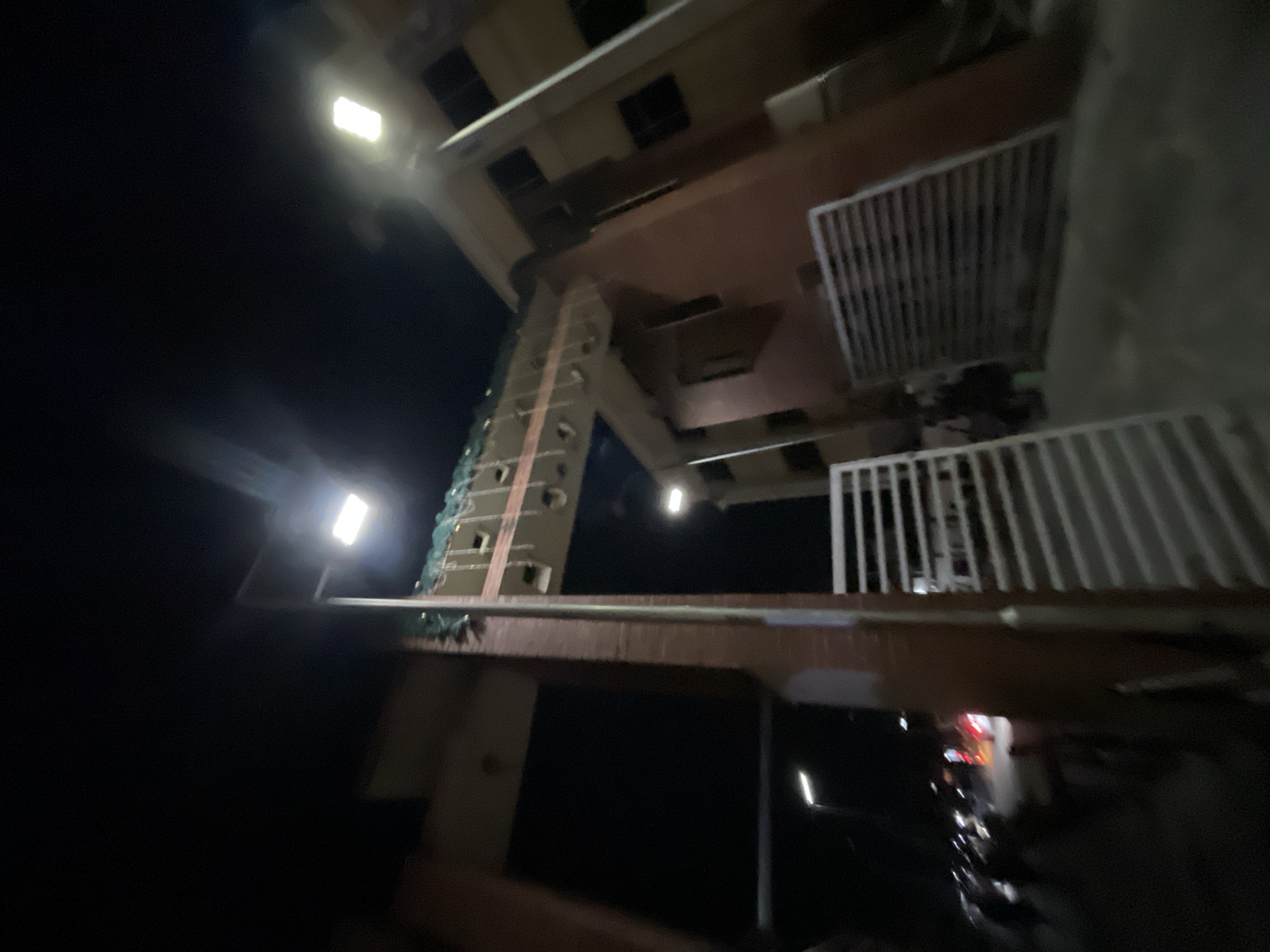The image, taken at night in Ado, Nigeria, shows a multistory building under construction, with a partially completed exterior. The structure is illuminated by two bright lights, one on the left and another in the middle. Scaffolding is visible, indicating ongoing construction work. The lower part of the image is obscured by a dark foreground, with hints of a reflective surface. The overall environment suggests an urban setting at night.