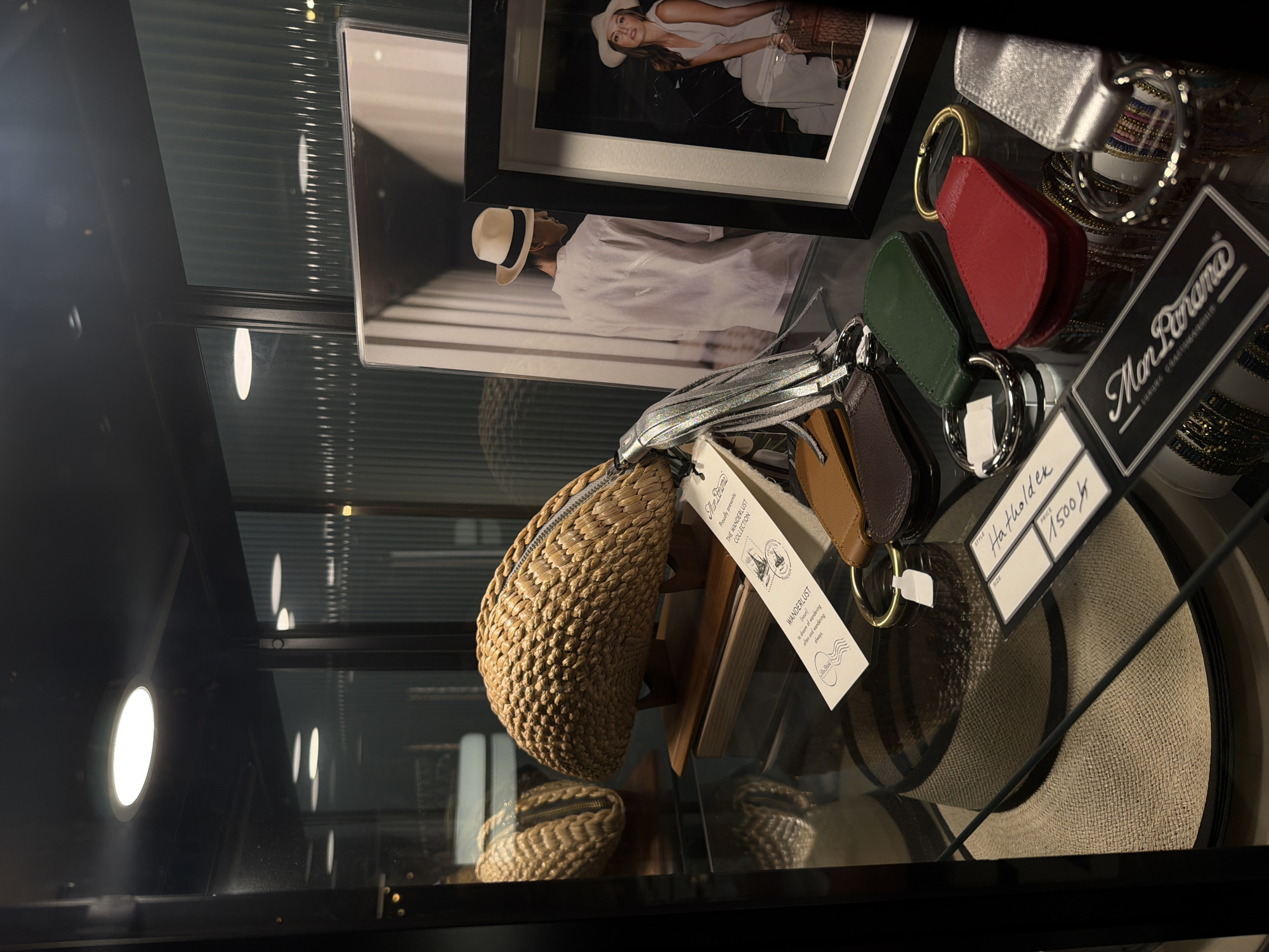 The image displays a retail display case, likely in a boutique or store, showcasing various accessories. In the background, a framed photograph features a woman in a wide-brimmed hat, possibly a fashion advertisement. Below the photo, a mannequin or display figure is shown wearing a similar hat and white clothing.
The foreground is filled with an assortment of items. A woven straw bag with a zipper is prominently displayed, with a tag that reads "THE WANDERLUST COLLECTION" and "WANDERLUST to dream of wandering often and wondering always" along with a logo that appears to be "Man Panana". Beside it, several small leather pouches in red, green, and brown, each attached to a metal ring, are also visible. A silver metallic accessory, possibly a small wallet or case, is also part of the collection.
A large straw hat with a dark band lies at the bottom of the display, positioned on a circular base. A tag attached to the hat states "STYLE Hatholdek", "SIZE", and "PRICE 1500". The overall scene suggests a display of luxury accessories, with a focus on hats and travel-themed items. The lighting in the display case creates reflections on the glass, and the background appears to be a dark, slatted wall, suggesting an indoor setting. There are no clear indications of the time of day or weather. The location context provided is Bangkok, Thailand.