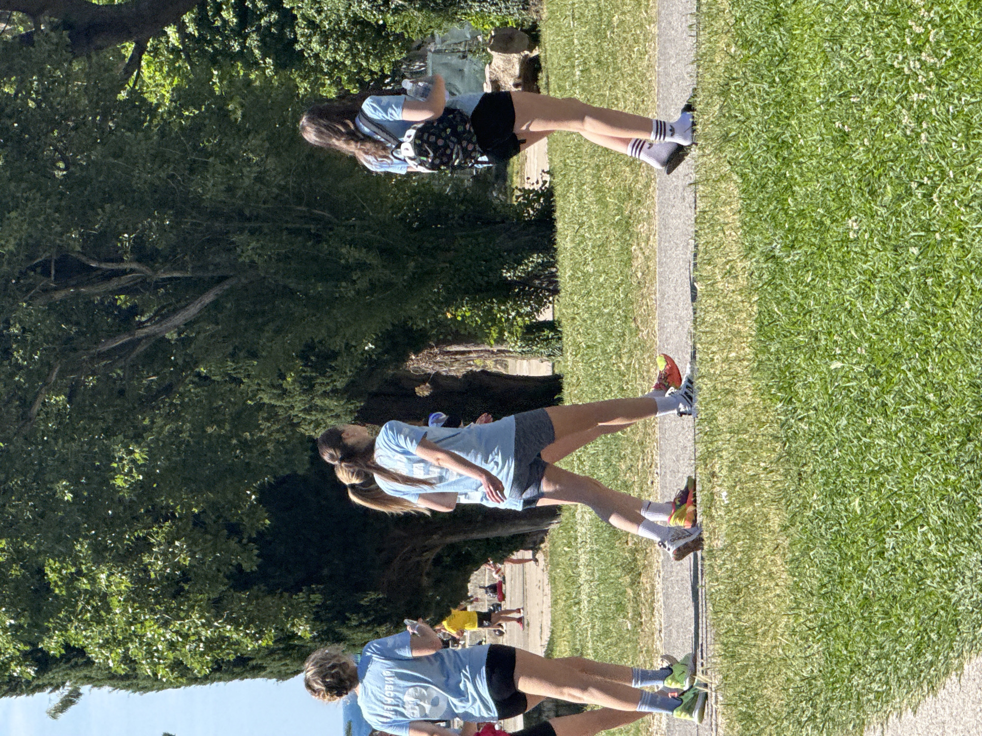 Three females, likely adolescents, are walking along a paved pathway. All are wearing light blue t-shirts and athletic shoes. The first female has dark hair and is carrying a backpack. The second female has her hair in a ponytail. There is greenery and trees visible on the left side of the image. The right side shows a grassy area. Other people are visible in the background, walking in the same direction. The location is speculated to be in the city of Budva, Montenegro.