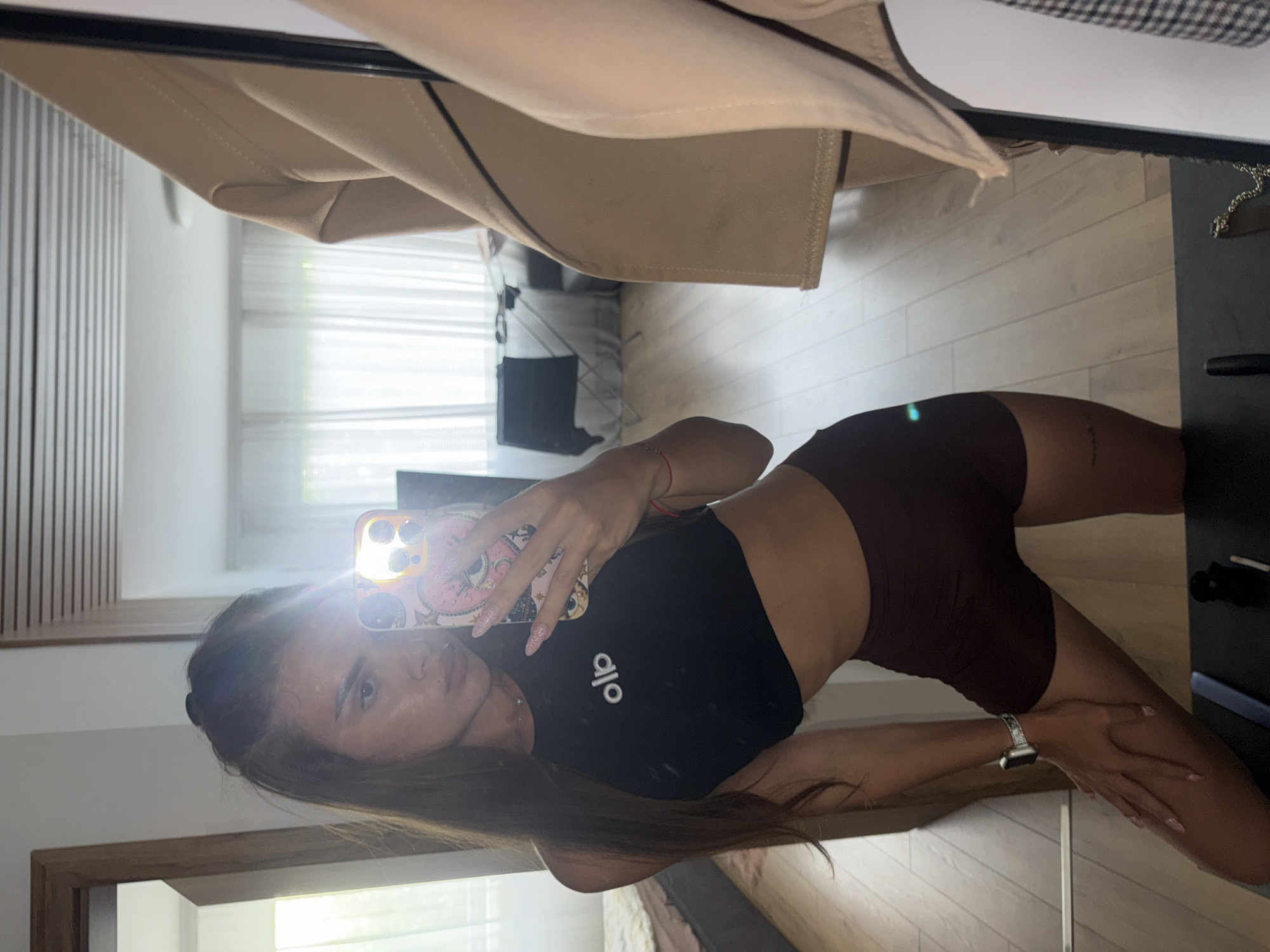A young woman is captured in a mirror selfie, positioned horizontally but depicting a vertical scene. She stands in an indoor room with light wooden floors and large windows covered by white blinds or sheer curtains, allowing bright natural light to fill the space. A wooden slatted wall panel is visible on the left.
The woman has long, dark brown hair and a neutral expression, looking directly at her phone. She is wearing a black crop top with the white text "alo" emblazoned on the front, and dark brown or black fitted shorts. An Apple Watch is on her left wrist. She holds a smartphone with a light pink, patterned case, which reflects a bright lens flare from the window. A light-colored coat or garment hangs above, partially obscuring the top of the mirror. To her right, a dark table or surface is visible. The overall setting suggests a modern and well-lit interior, likely a bedroom or living area, during the daytime.