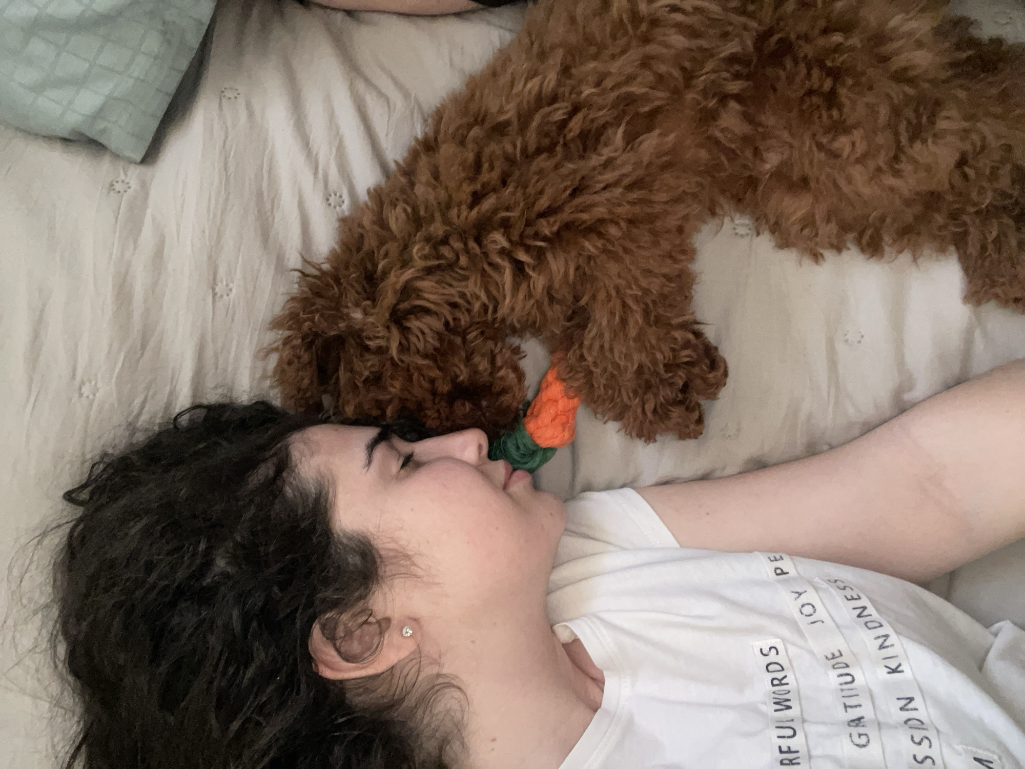 A woman with dark, curly hair lies on her back on a bed, with a brown, curly-haired dog on top of her. The dog's head is near her face, and it is holding a toy carrot in its mouth. The woman is wearing a white t-shirt with words printed on it. A pillow is visible in the upper left corner of the frame. The bed has a light-colored patterned sheet. There is no indication of the city, Sofia, Bulgaria, in the image.