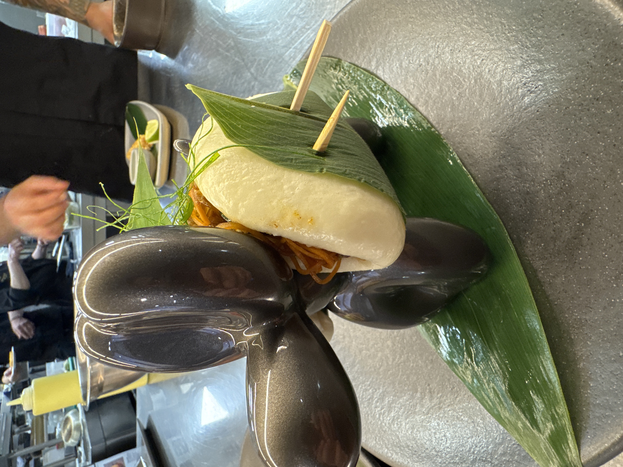 The image shows a close-up of a stylized, dark grey, balloon-dog-shaped sculpture presenting a bao bun. The bao bun is wrapped in a large green leaf and secured with two wooden skewers. The bun is filled with shredded orange material, possibly carrots or pork. A few sprigs of green garnish are also visible. The sculpture and bun are placed on a dark grey plate with a textured surface, and underneath the bun is a long, glossy green leaf. In the background, a kitchen environment is visible, with stainless steel surfaces, containers, and people in dark chef uniforms. One person's hands are in the frame, seemingly in the process of preparing food. The scene is well-lit, with reflections visible on the sculpture and the kitchen surfaces. There is no indication of the city of Sofia, Bulgaria in the image.