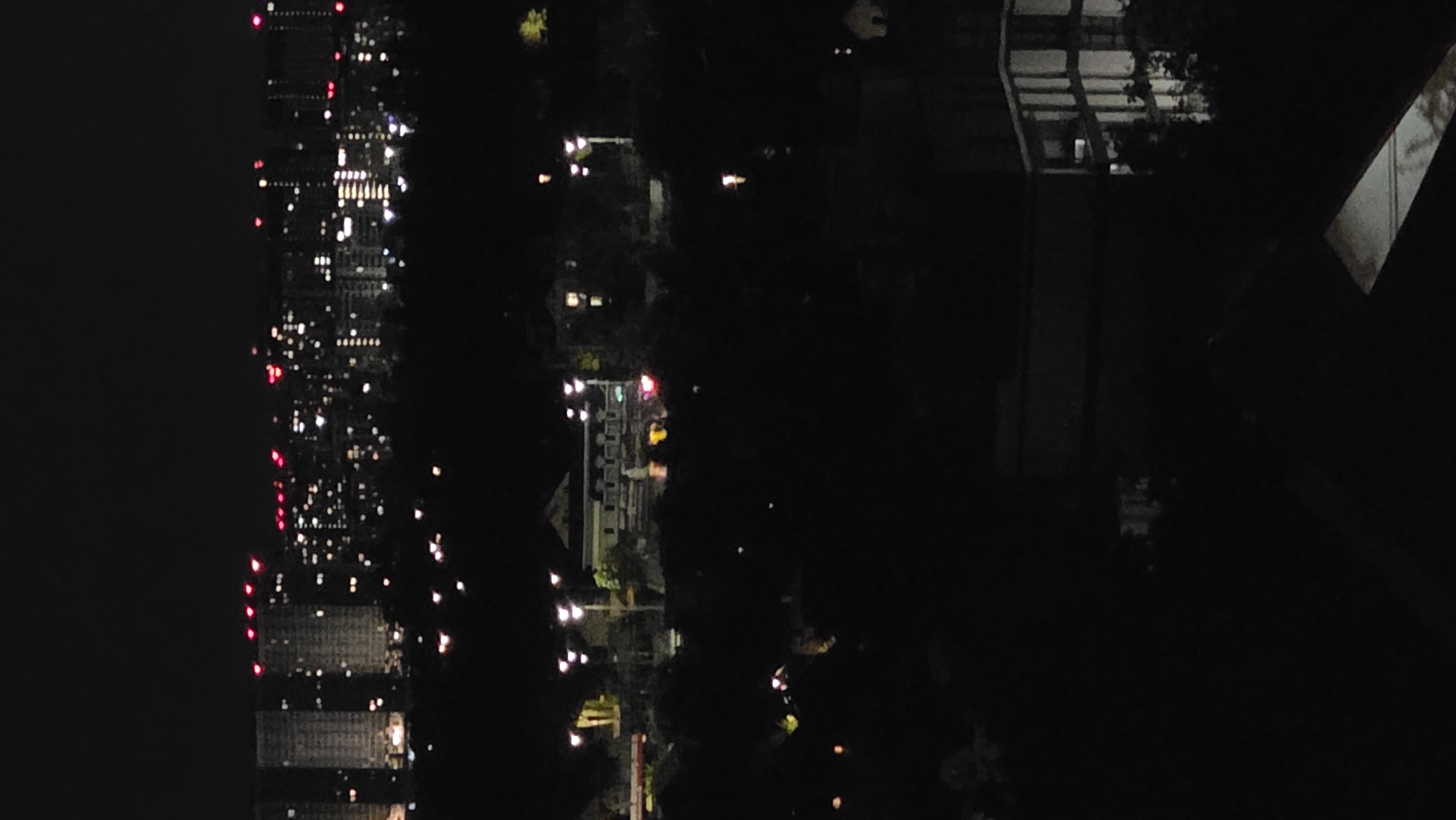 A nighttime, long-shot, vertically oriented, eye-level photograph depicts a city skyline with a dark, tree-filled foreground and midground. The background shows a blurred city skyline with numerous lights. Several tall buildings with illuminated windows are visible. Some of the buildings have red lights on their upper levels. The midground features a more distinct building with a light-colored facade and a patterned roof, surrounded by trees and streetlights. The foreground is almost entirely dark, with a hint of a building structure on the right side of the frame, appearing to be a modern, multi-story building with illuminated windows.