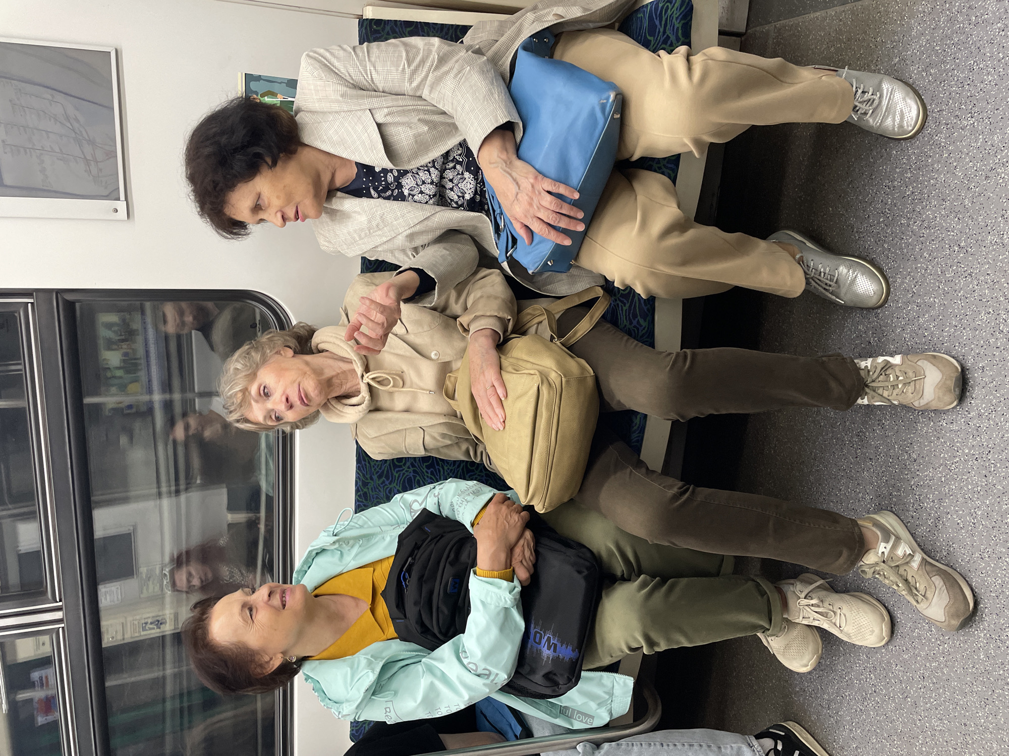Three elderly women are sitting on a bench in what appears to be a subway or train car. The woman on the left is wearing a light blue jacket over a yellow shirt and is holding a black backpack. The woman in the middle is wearing a beige hooded jacket and has a beige shoulder bag. The woman on the right is wearing a patterned jacket over a dark blue shirt and has a bright blue handbag on her lap. All three women are looking in different directions, and their expressions suggest they are engaged in conversation. The environment is utilitarian, with a patterned bench and gray flooring. There are reflections in the window behind them, showing other passengers and the interior of the train. The overall scene depicts a moment of everyday life on public transport.