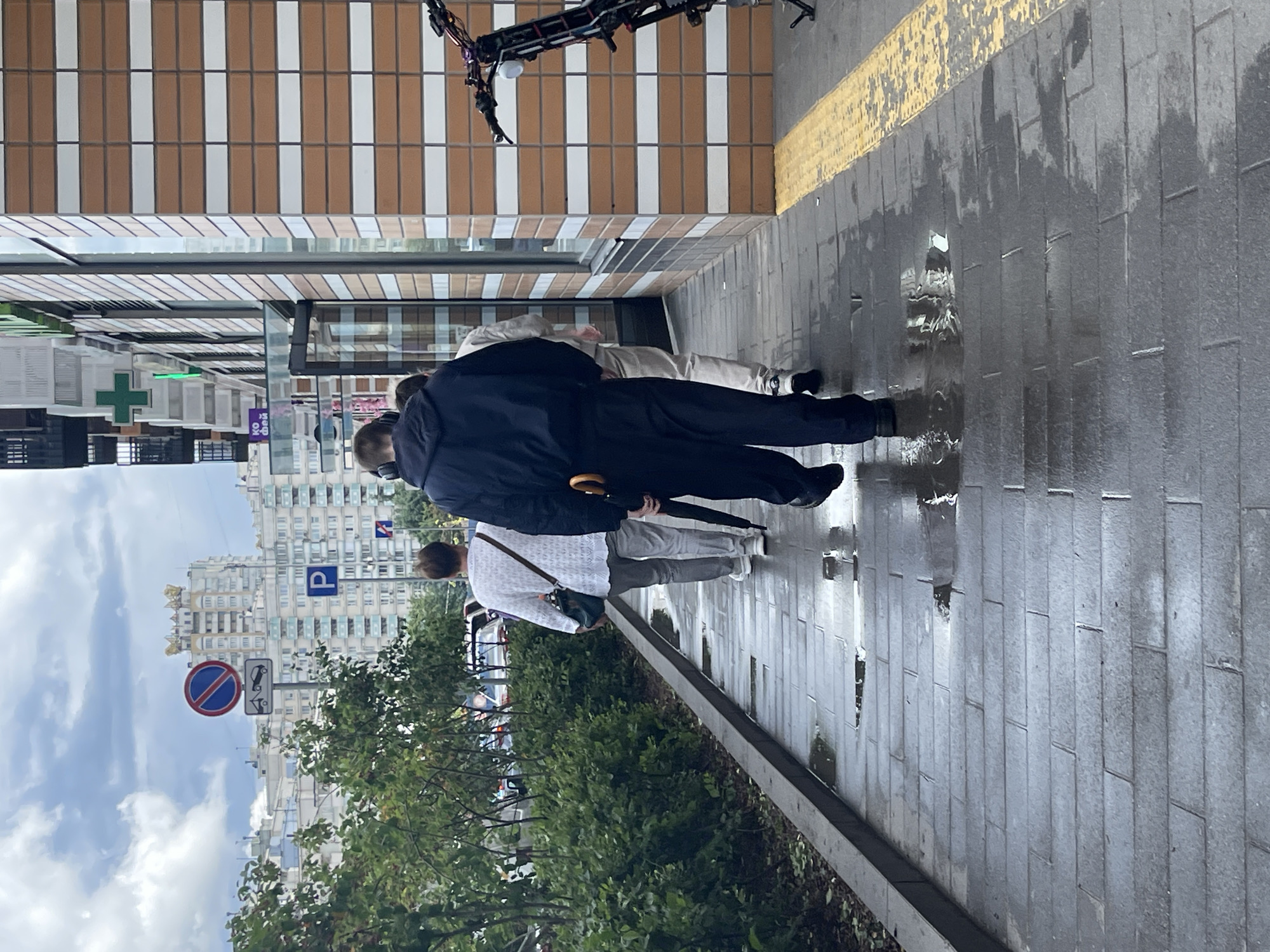 The image depicts a street scene in Moscow, Russia, on a cloudy day with wet pavement indicating recent rain. Three individuals are walking away from the camera on a tiled sidewalk. The person in the foreground is wearing a dark blue hooded jacket and dark pants, carrying an umbrella with an orange handle. To their left, a person in a white patterned sweater and grey pants walks ahead, with a dark green bag slung over their shoulder. A third person, partially visible, walks behind the first individual.
To the left of the sidewalk is a green bush and trees, with a street and cars visible in the background. On the right side of the image is a building with a facade of alternating brown and white tiles, featuring large windows. A green cross sign, likely for a pharmacy, is visible on the building. In the distance, several multi-story apartment buildings are visible under a grey, cloudy sky. A bicycle or scooter is parked on the right side of the image, near the building. Traffic signs, including a "no stopping" sign and a parking sign, are visible in the background.