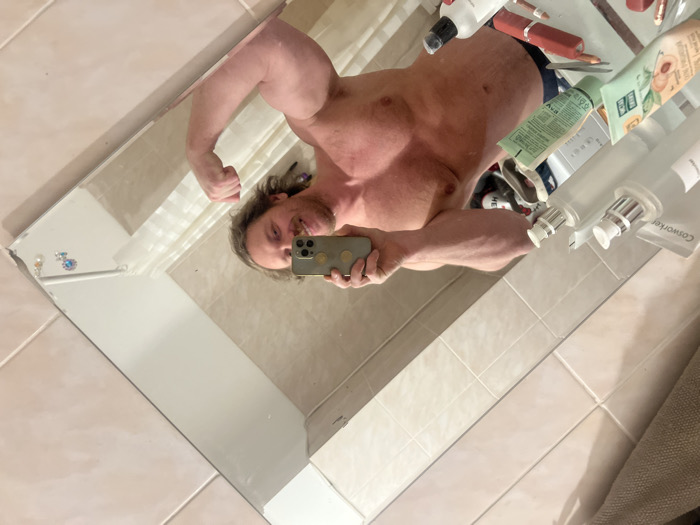 A muscular man poses for a selfie in a bathroom mirror, flexing his left bicep. The man is shirtless, revealing a toned physique. The mirror reflects a cluttered shelf with various cosmetic products. The bathroom features light-colored tile and a neutral-toned shower curtain or towel visible in the corner of the reflection. The overall impression is one of self-admiration in a domestic setting.