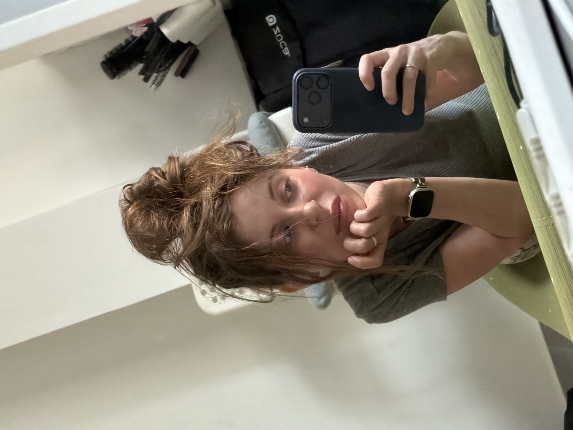 A selfie captured by a woman with light skin and brown hair styled in a messy bun. She's wearing a grey shirt and a smart watch on her left wrist, holding a dark blue smartphone to take the photo. In the background, there is a white wall, a shelf with hairbrushes, and a black bag. The location is likely indoors, but there is no indication of it being in Bat Yam, Israel. The scene focuses primarily on the woman taking a selfie.