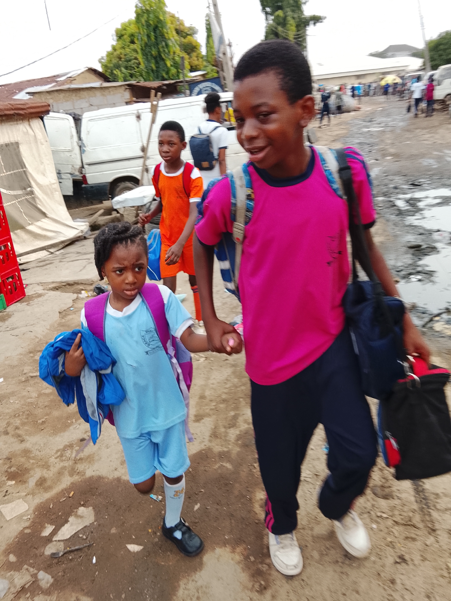 A group of three children walk together on a dirt road with puddles of water in what appears to be Abuja, Nigeria. In the foreground, a young girl wearing a light blue school uniform with matching shorts and socks with black Mary Jane shoes. She carries a dark blue jacket in her left hand and wears a purple backpack. She is holding hands with a boy to her right who wears a pink shirt, navy pants with pink stripes, and white sneakers. He carries a black bag in his right hand and also wears a backpack. Behind them and to the left is another boy wearing an orange shirt and shorts with soccer socks. In the background, there are white vans parked next to a building with a tan tarp, and blurred figures of other people walking in the distance.