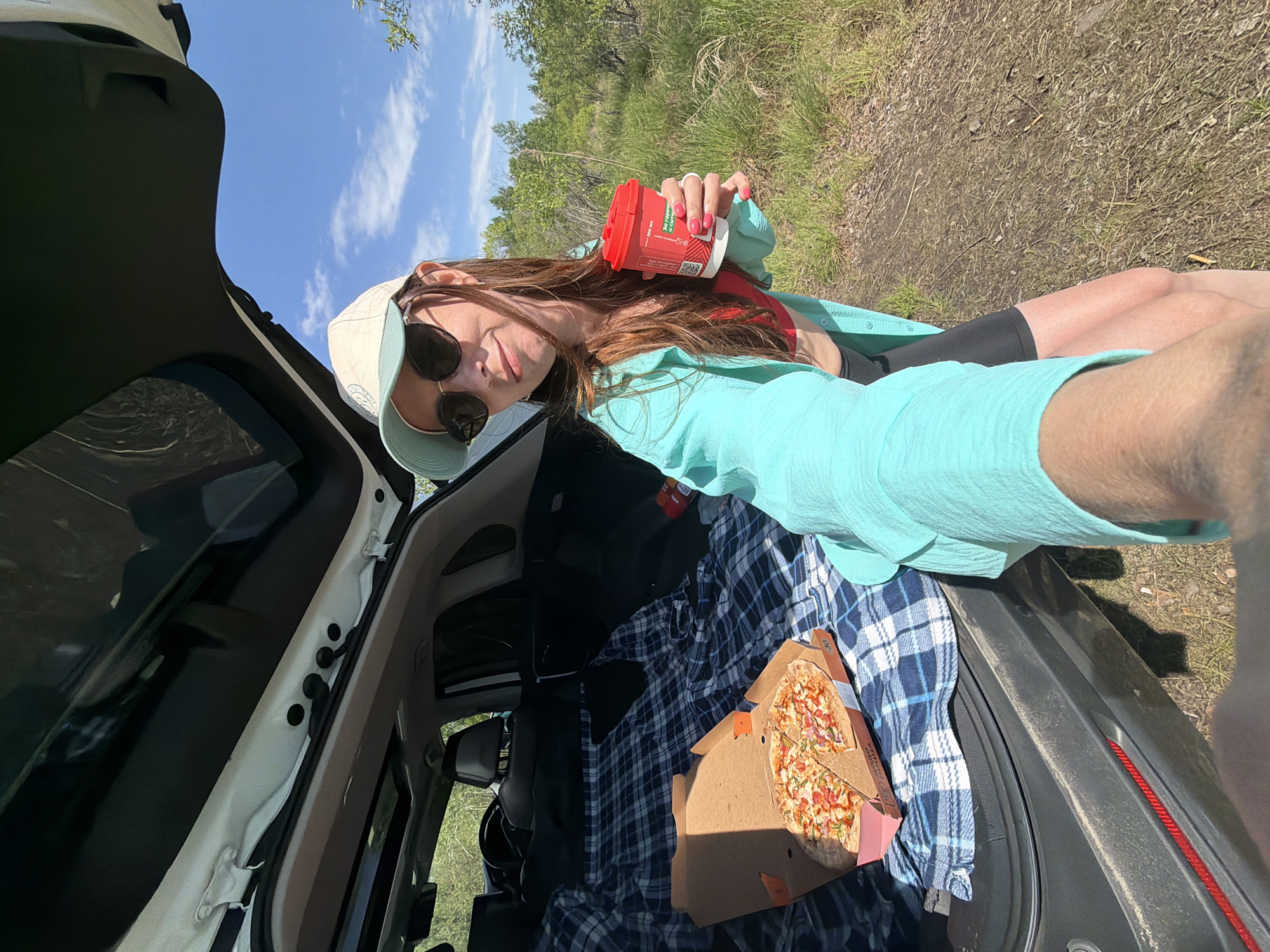 A woman wearing a light green cap, round sunglasses, a red top, black shorts, and a light blue button-down shirt is holding a red coffee cup in her right hand. She is standing by the open trunk of a gray SUV. Inside the trunk, on top of a blue and white plaid blanket, is an open pizza box with a pizza inside. The background shows a grassy area with trees and a blue sky. The location is not specified in the image.