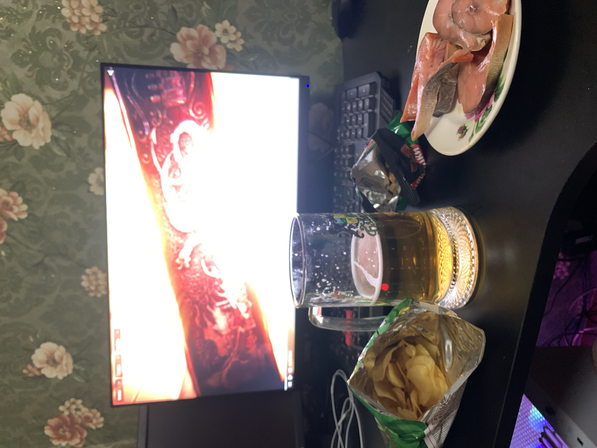 The image shows a close-up of a table with food and drink in front of a computer monitor. On the table, there is a glass of beer, a bag of potato chips, a plate of raw fish slices, and what appears to be a bag of dried fish snacks. The computer monitor displays a bright, abstract image with red and yellow tones. The background wallpaper has a floral pattern in green and beige. The scene appears to be set in a domestic environment, likely a home, and there is no indication of the location being in the city of Desyatikhatka, Russia. No people are visible in the image.