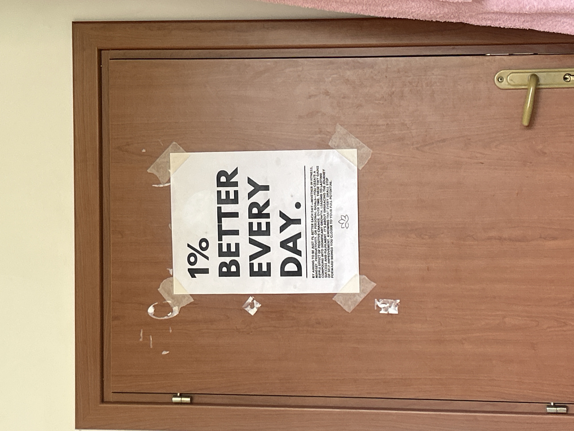 The image shows a wooden cabinet door with a piece of paper taped to it. The paper has the text "1% BETTER EVERY DAY." printed on it in large, bold letters. Below the text is a smaller paragraph describing the concept of incremental improvement. There are pieces of masking tape securing the paper to the door in each corner and in the middle along the top and bottom edges. There is a metal handle on the right side of the door. The door is part of a built-in cabinet or cupboard with a wooden frame. A pink item of clothing (possibly a towel) rests on top of the cabinet door. The wall to the left of the door is off-white. The image was not taken in Veliko Tarnovo, Bulgaria, as the query requested, because the image does not contain sufficient data to identify the location.
