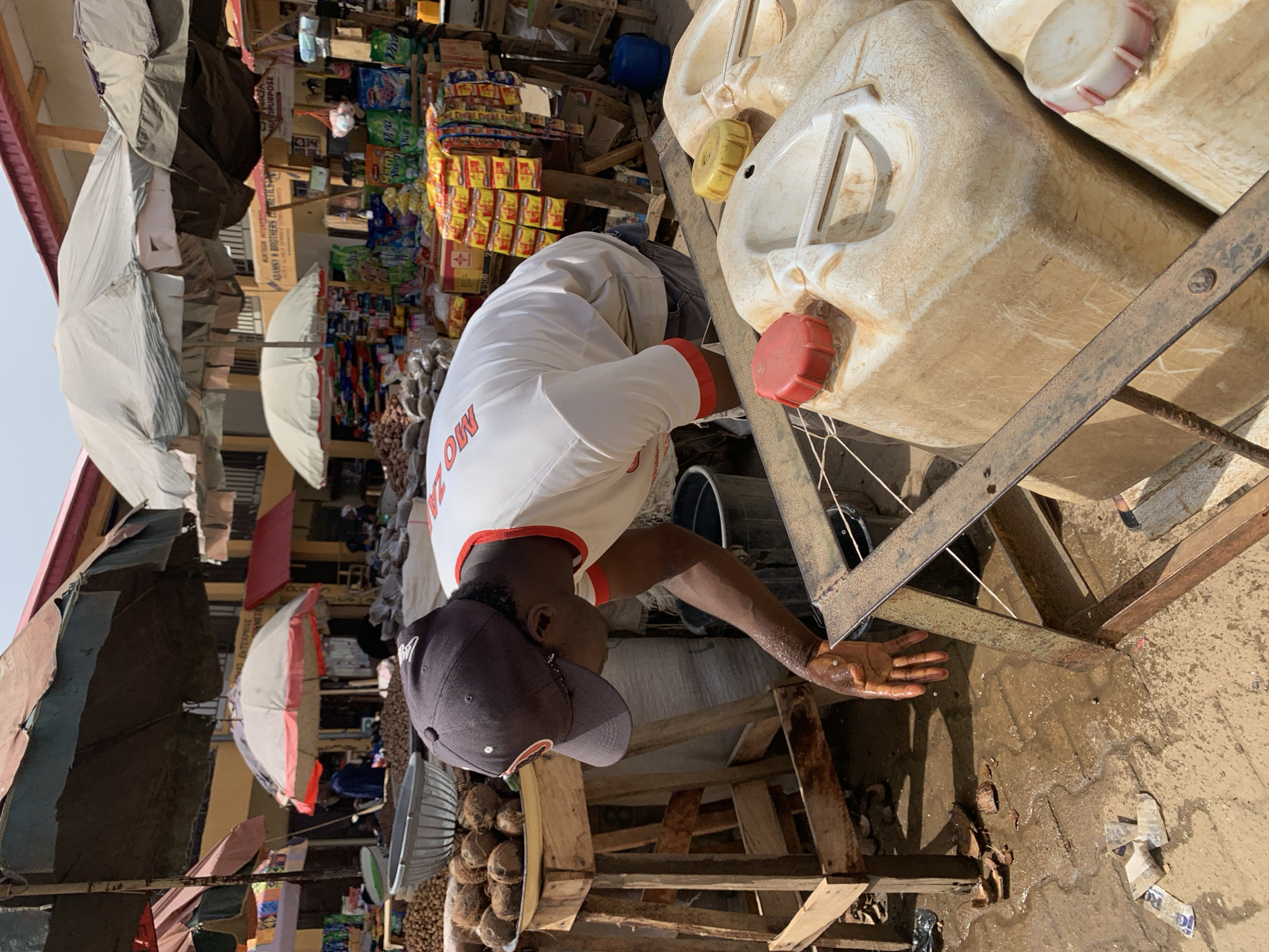 This image presents a dynamic outdoor market scene in Kafanchan B, Nigeria, captured during bright daylight hours. The primary subject is a man, seen in profile, wearing a white short-sleeved shirt with red trim, a dark baseball cap, and dark trousers. He is bent over, his hands wet, appearing to handle liquid from large, light-colored plastic jerry cans that rest on a rusty metal frame. The ground around him is wet with puddles, and some discarded paper and debris are visible.
The background reveals a vibrant marketplace filled with various stalls. These stalls are packed with an assortment of packaged goods, including snacks, detergents, and other household products. Overhead, multiple umbrellas and makeshift canopies, some white and others multicolored, provide shade from the sun. Wooden crates and pallets serve as display stands and support structures for the merchandise.
Notable details include a prominent yellow sign in the mid-ground that reads "ADAMS & BROTHERS RETAILS ENTERPRISE," indicating a local business. Other visible text includes "MULTPURPOSE," "DETERGENT," and "VIVA" on various product packages. A partial word, "MO," can be seen on the man's shirt. Near the man's feet, a metal bowl holds small round items, possibly nuts or seeds, adding to the market ambiance. The scene depicts a bustling commercial environment, typical of an African open-air market, focused on daily trade and necessities.