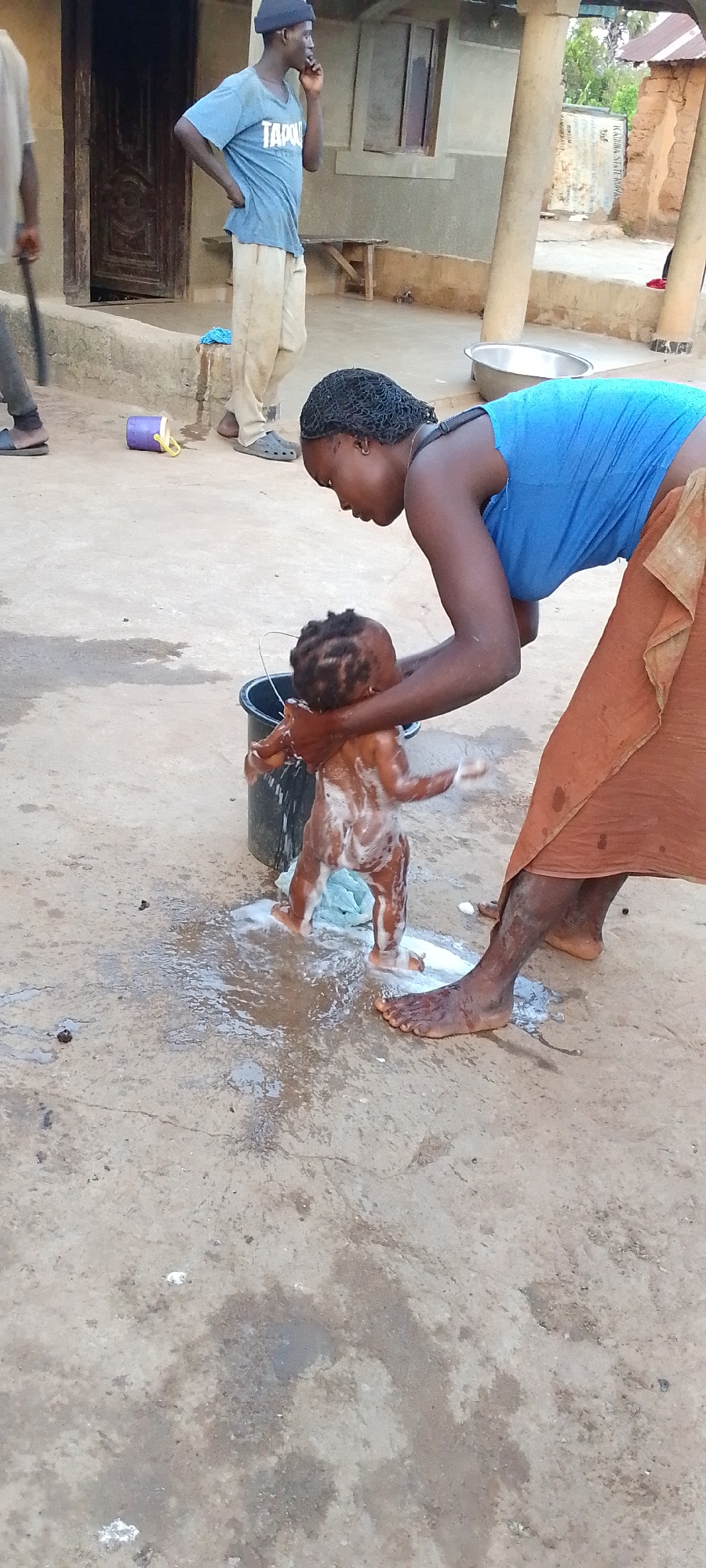 The image depicts an outdoor scene in Kamantan, Nigeria, likely during daylight hours. The primary activity is a woman bathing a young child. The woman, wearing a blue top and an orange cloth, is leaning over a black bucket, gently washing the soapy child. The child is covered in suds and appears to be standing, with hands raised. Water and soap are splashed on the concrete ground around them.
In the background, a man wearing a blue t-shirt with the word "TAPOLI" visible, light-colored pants, and a dark beanie stands with his hand on his hip, looking away from the camera. Further back, parts of a building with wooden doors and windows are visible, along with pillars and what appears to be dry vegetation in the distance. A small purple and yellow bucket lies on the ground near the man. The overall setting suggests a rural or semi-rural environment. The ground is uneven concrete, with some cracks and scattered debris. The atmosphere appears casual and domestic.
