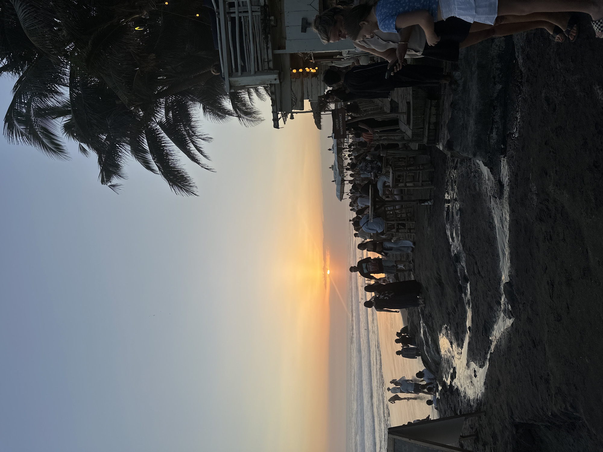 This image depicts a crowded beach scene at sunset. The sky is a gradient of soft blues and oranges, with the sun low on the horizon, casting a warm glow over the ocean. Waves are gently rolling onto a dark sand beach. In the foreground, a small stream of water snakes across the sand.
Numerous people are scattered across the beach and in the shallow water. Some are standing, some are sitting, and others are walking. Towards the right side of the frame, a cluster of people are gathered around what appears to be a raised wooden structure, possibly a restaurant or bar, with thatched umbrellas in the background. Palm trees are visible along the edge of the structure, their fronds silhouetted against the sky. String lights are hung from the structure, adding to the ambiance.
In the middle ground, a group of people are walking along the shoreline. A few individuals are also seen in the water. In the far distance, more people are visible on the beach and in the ocean. The overall atmosphere appears to be lively and social, with many people enjoying the sunset.