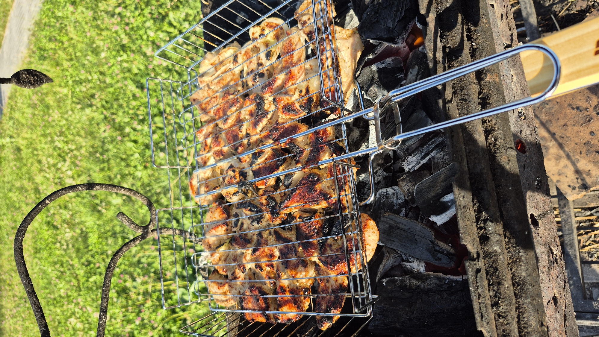 A close-up, high-angle shot captures a metal grilling basket filled with pieces of chicken being cooked over hot coals. The chicken pieces, arranged in rows within the basket, display char marks and varying degrees of browning from the grill. The grilling basket rests on a bed of dark charcoal, with some glowing embers visible beneath. The scene is set outdoors with a blurred background of green grass and small pink wildflowers, suggesting a park or garden setting. A black, curved metal stand is visible in the upper left portion of the frame, and a wooden handle extends into the lower right corner. The lighting suggests it is daytime, with sunlight casting shadows. There are no people or animals present.