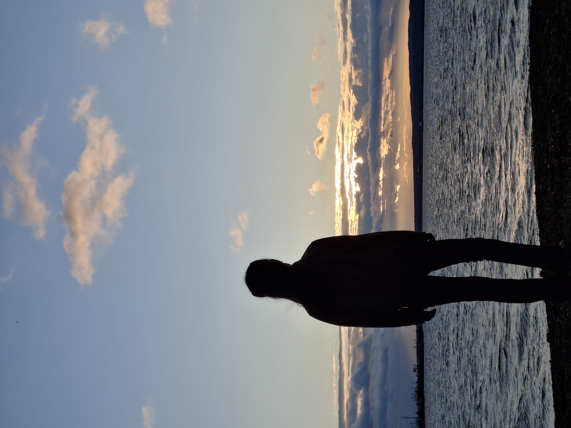 A lone figure stands silhouetted against a sunset over the Kama River in Chaikovsky, Russia. The individual, facing away from the camera, has their back to the viewer, with long hair pulled back. They are wearing dark pants and a jacket. The sky is a gradient of blue and orange, with scattered clouds illuminated by the setting sun. The river water is choppy, reflecting the colors of the sky. In the distance, along the horizon, is a dark silhouette of the land, with hints of industrial structures on the left side of the image. The foreground consists of dark pebbles on the riverbank.