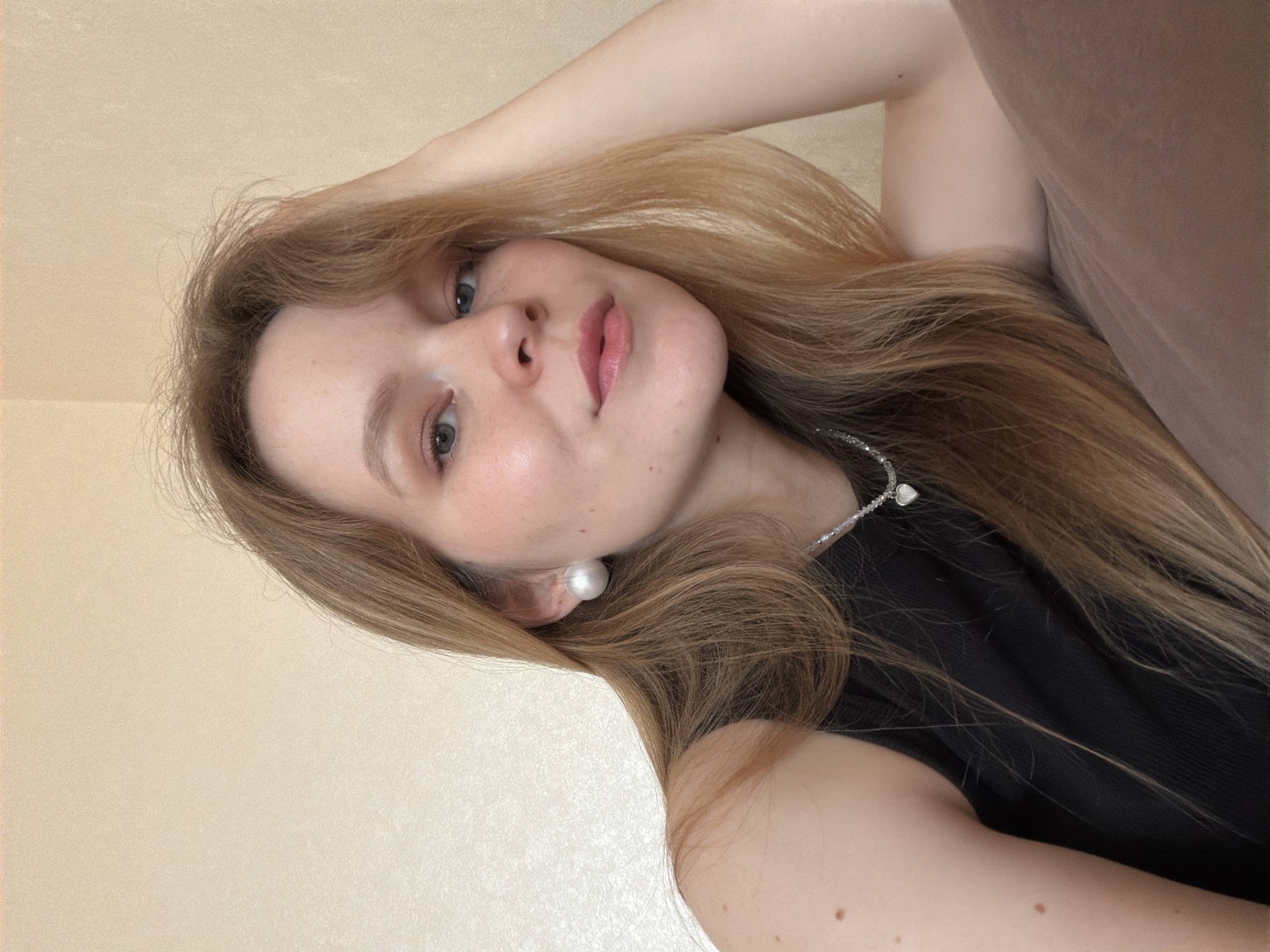 The image is a close-up, waist-up, selfie-style portrait of a young Caucasian woman with long, light brown hair, pale skin, and light blue eyes. She is wearing a black sleeveless top, a pearl stud earring in her left ear, and a silver necklace with a small heart pendant. Her left arm is bent, with her hand resting on her head. Her right arm is partially visible, resting on a muted brown surface, likely a piece of furniture. The background is a plain, light beige wall with a subtle textured finish. The lighting appears to be soft, coming from the front and slightly from the left, creating a gentle illumination on her face. There is no discernible indication of the location being Kazan, Russia, or any specific urban environment. The scene is indoors and appears to be a personal, casual photograph.