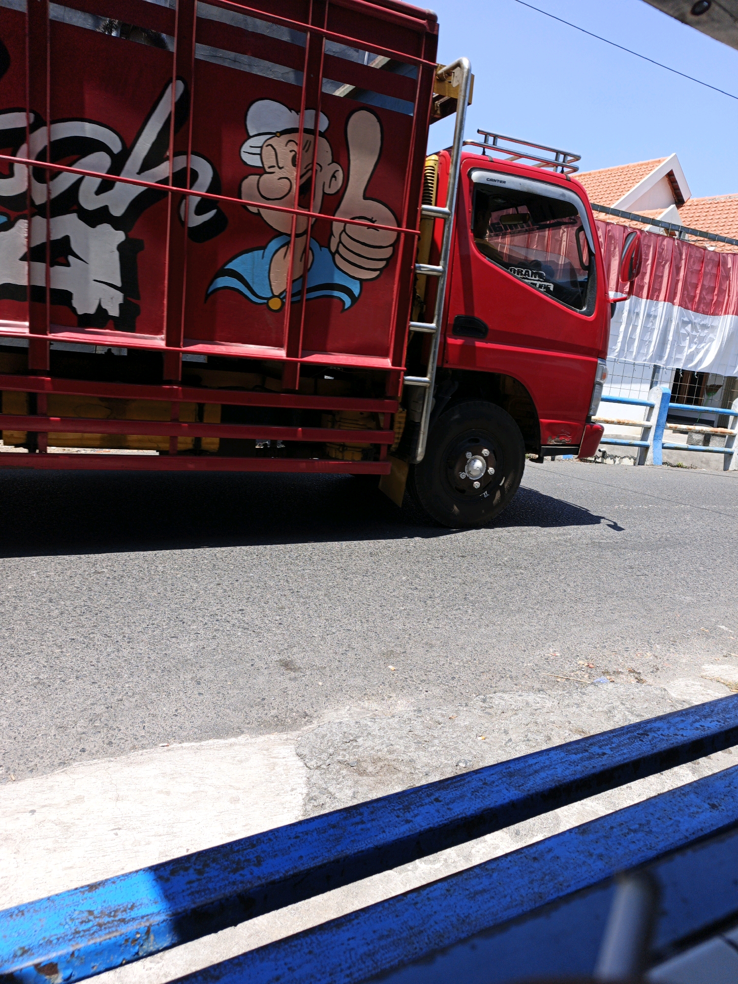 The image captures a red truck with a cartoon character, Popeye, painted on its side, parked on a street in Ketegan, Indonesia. The truck is adorned with red and white striped banners, suggesting a festive or promotional event. The street appears to be paved with asphalt, and the surrounding environment includes buildings with tiled roofs, a blue and white fence, and a concrete sidewalk. In the foreground, a blue bench made of metal bars is visible, with part of the truck's shadow cast upon it. The sky is clear and blue, indicating a sunny day.