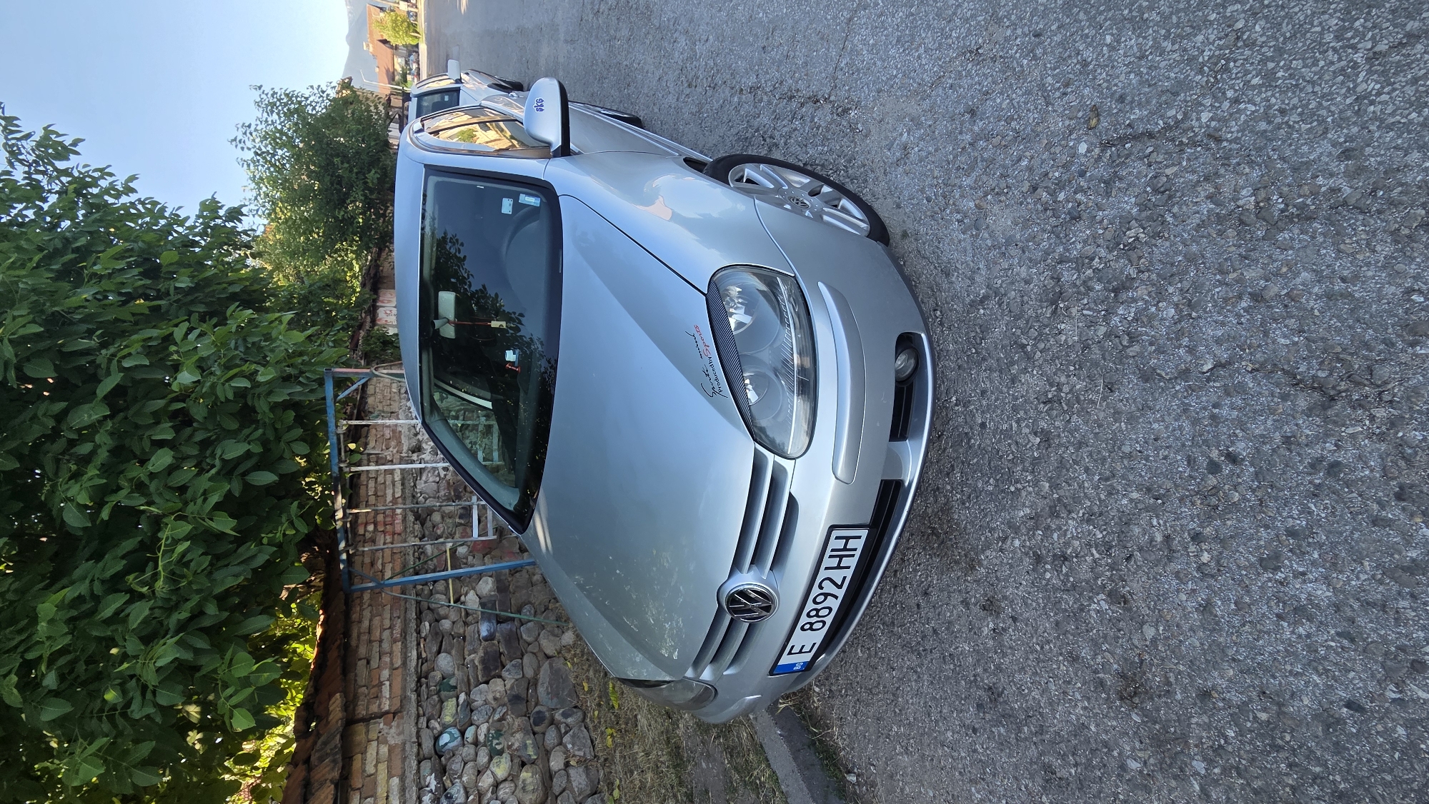 A silver Volkswagen Golf is parked on an asphalt street in the city of Bania, Bulgaria. The car is facing the viewer's right and is partially obscured by a stone wall and a large tree with green foliage. A metal swing set is visible behind the car and to the left. The car has a Bulgarian license plate that reads "E 8892 HH". There is a sticker on the hood of the car that says "Sports and produced by Sports". The sky is clear and blue. In the background, there are other cars parked on the street and buildings.