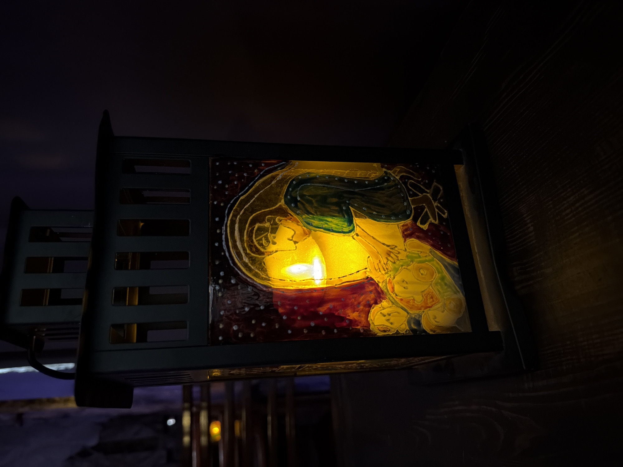 The image features a decorative, illuminated wall-mounted lantern. The lantern has a dark metal frame with horizontal bars and a rectangular panel displaying an image of what appears to be a person and other figures, possibly mythological or allegorical. The panel is internally lit, casting a warm, yellowish glow. The background is dark, suggesting a dimly lit interior space. The wooden wall to the right is a dark brown color and displays noticeable grain. The photograph is taken at an upward angle, showing a partial view of what appears to be an interior, possibly located in Saint Petersburg, Russia.
