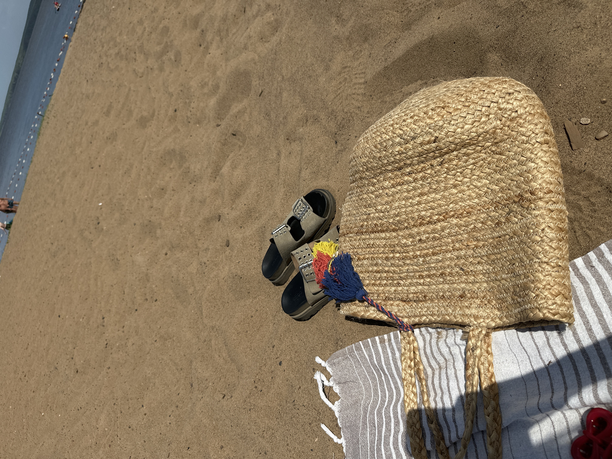 The image depicts a beach scene, likely on a sunny day. The beach is composed of sand, and in the upper left, the edge of a body of water with a line of buoys can be seen.
In the foreground, a beach bag made of woven material is resting on a striped towel. A pair of sandals are positioned next to the bag. The bag has a multi-colored tassel attached to it.
The image was likely taken in Chaikovsky, Russia, based on prior information.