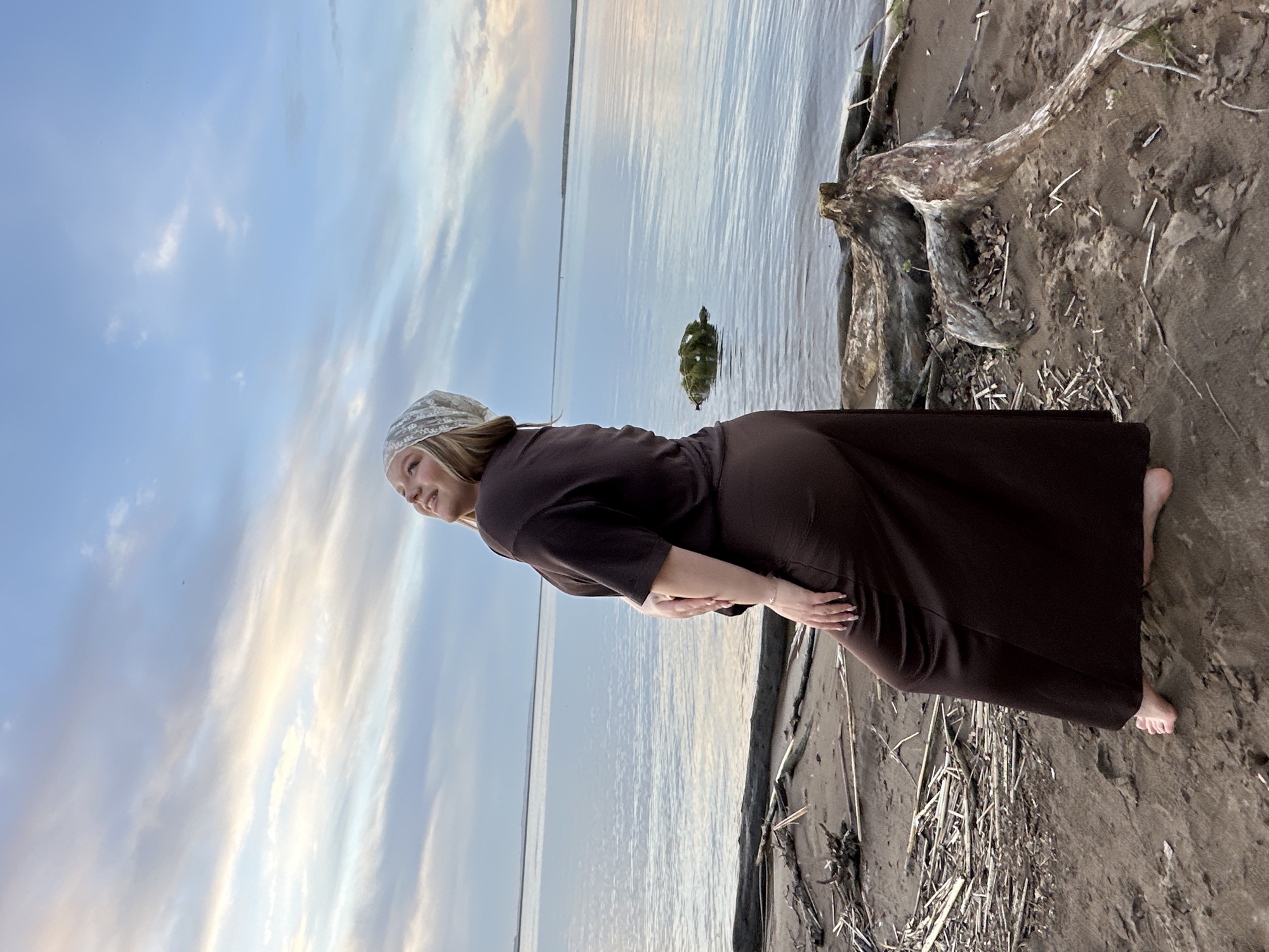 A woman stands on a beach in Chaikovsky, Russia, near the Kama River. She wears a long, dark brown dress and a white head covering. She gazes out toward the river. The sky is light blue with streaks of white clouds. The beach is sandy, with some driftwood and reeds. A small, rocky island is visible in the river.