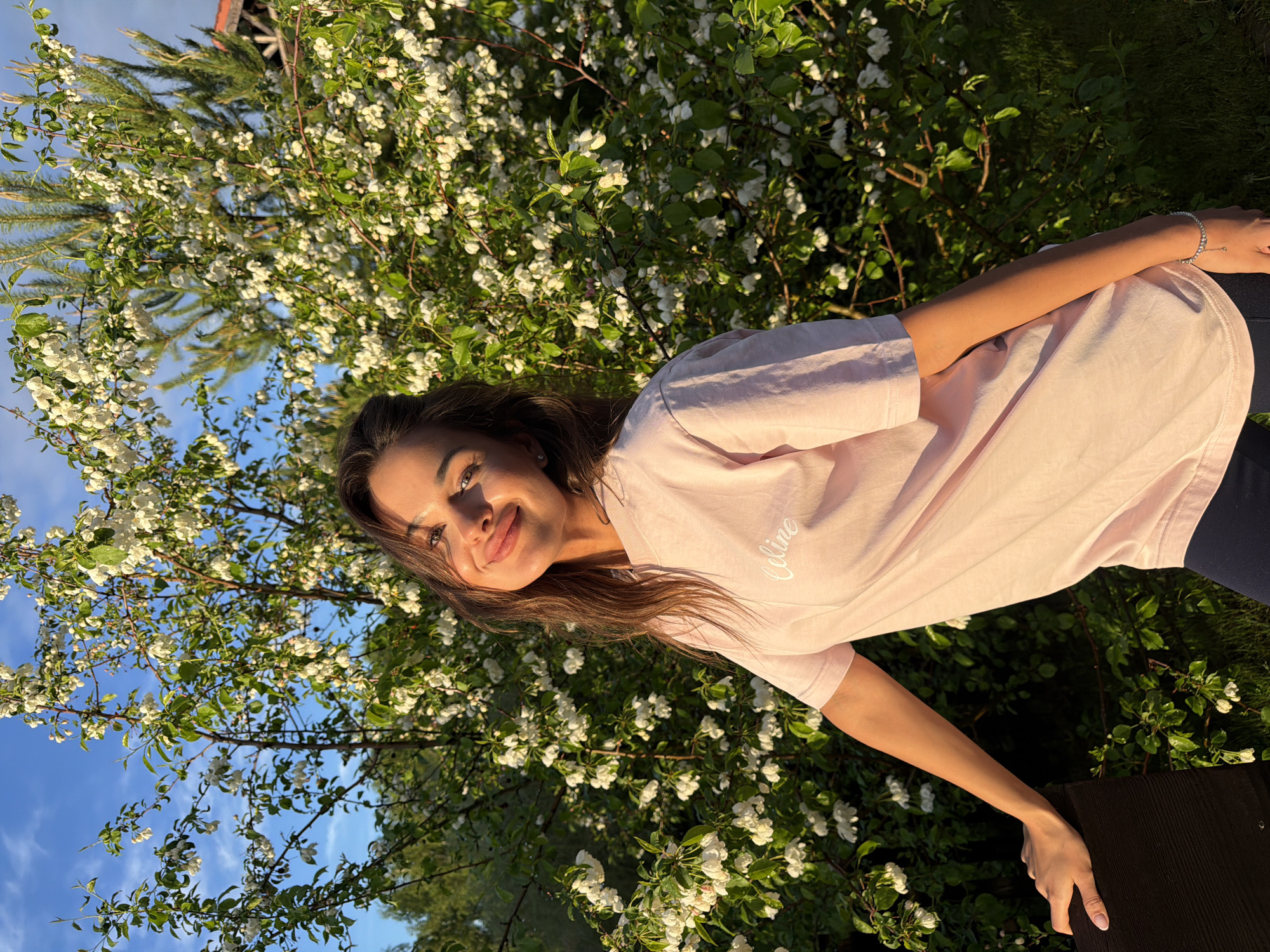 A young woman with long brown hair smiles at the camera, positioned in front of a blossoming tree. She wears a light pink t-shirt. The background includes parts of the tree with white flowers, a patch of blue sky, and a hint of a building structure. The scene appears to be outdoors, with natural light illuminating the subject and surroundings. The location is Shamoni, Russia.