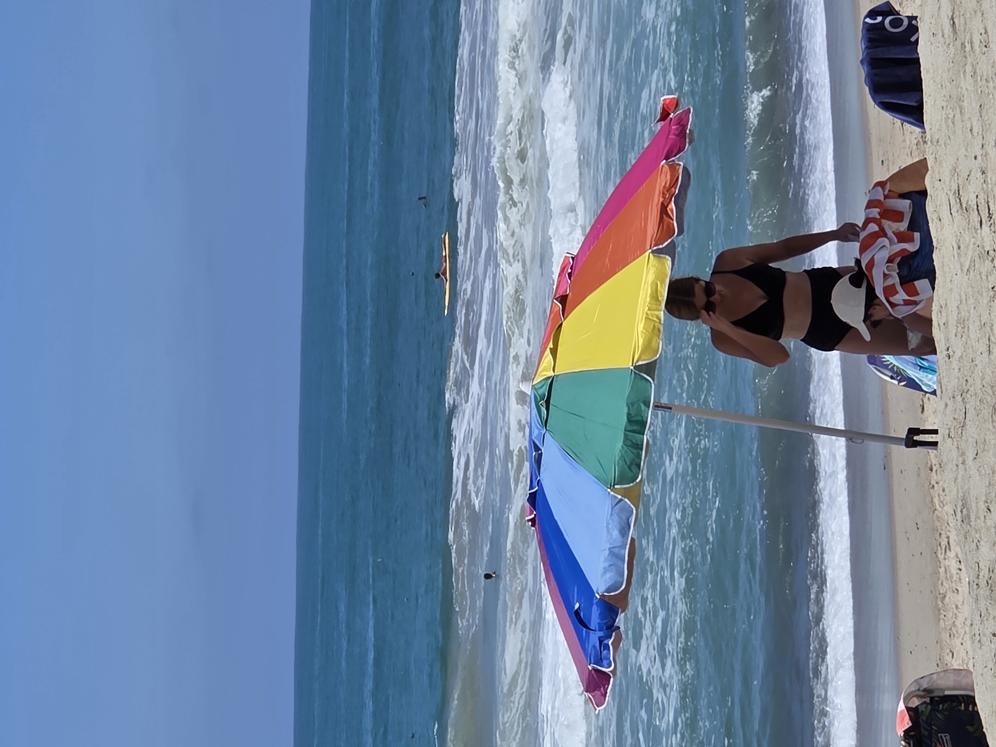 The image depicts a sunny day at the beach, likely in San Clemente, California, given the characteristic surf and sand. The foreground features a colorful, multi-panel beach umbrella providing shade. Beneath the umbrella, two individuals are present. A woman wearing a black bikini and sunglasses stands, adjusting her sunglasses. Next to her, a person wearing a white cap and a towel draped over their shoulders is positioned lower down on the sand. In the background, the ocean displays moderate waves and the horizon shows a clear blue sky. Several surfers can be seen in the water, with one person lying on a surfboard. The overall scene conveys a typical beach day with people enjoying recreational activities.