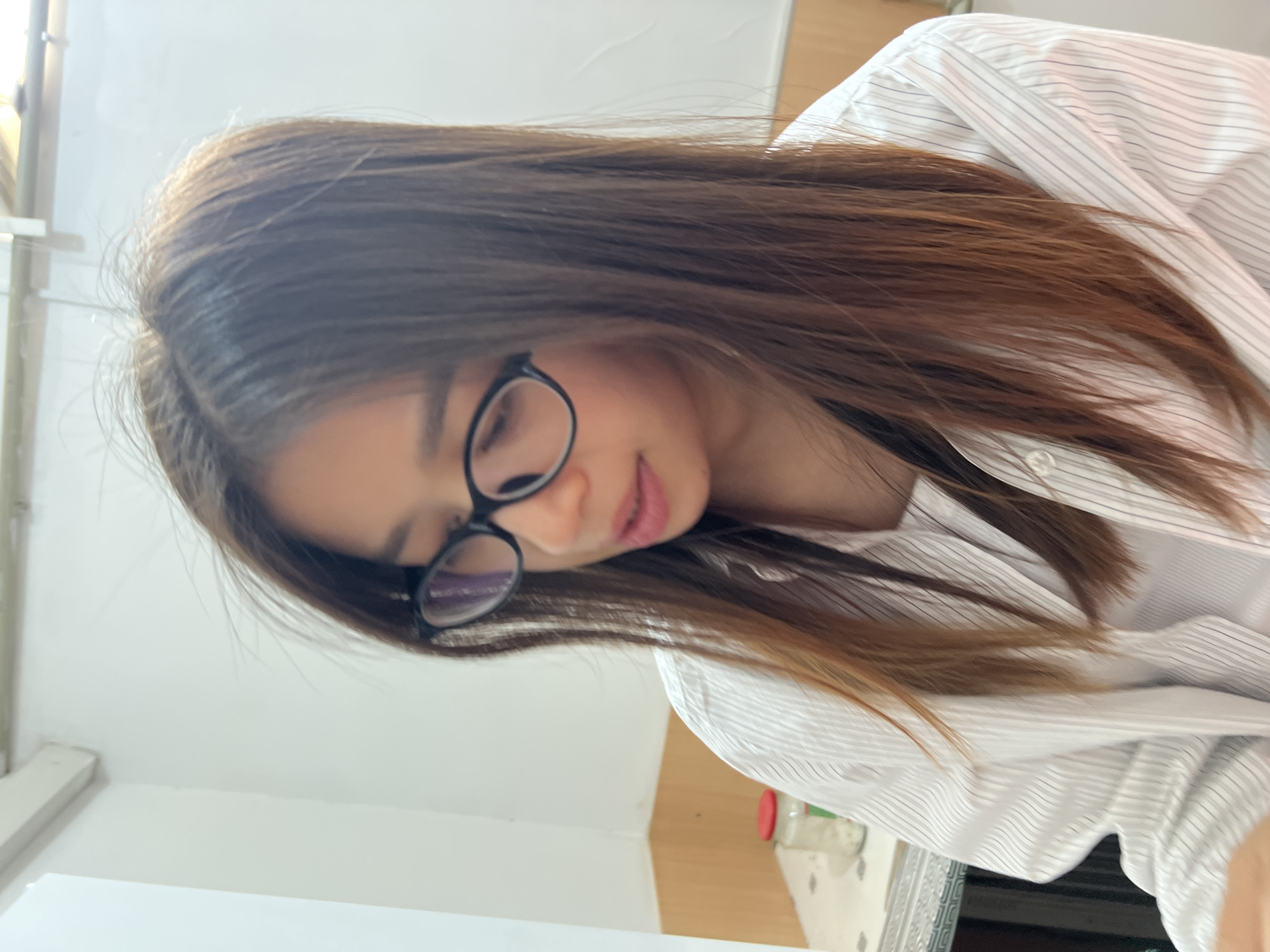 A young woman with long, straight brown hair and glasses is shown from the chest up. She is wearing a white button-down shirt with thin black pinstripes over a white tank top. The background appears to be an indoor setting with white walls and a beige tiled area in the lower left corner. A glass jar with a red lid is visible on a table in the background. The scene is likely in a domestic or casual environment within Kadamjay, Kyrgyzstan.
