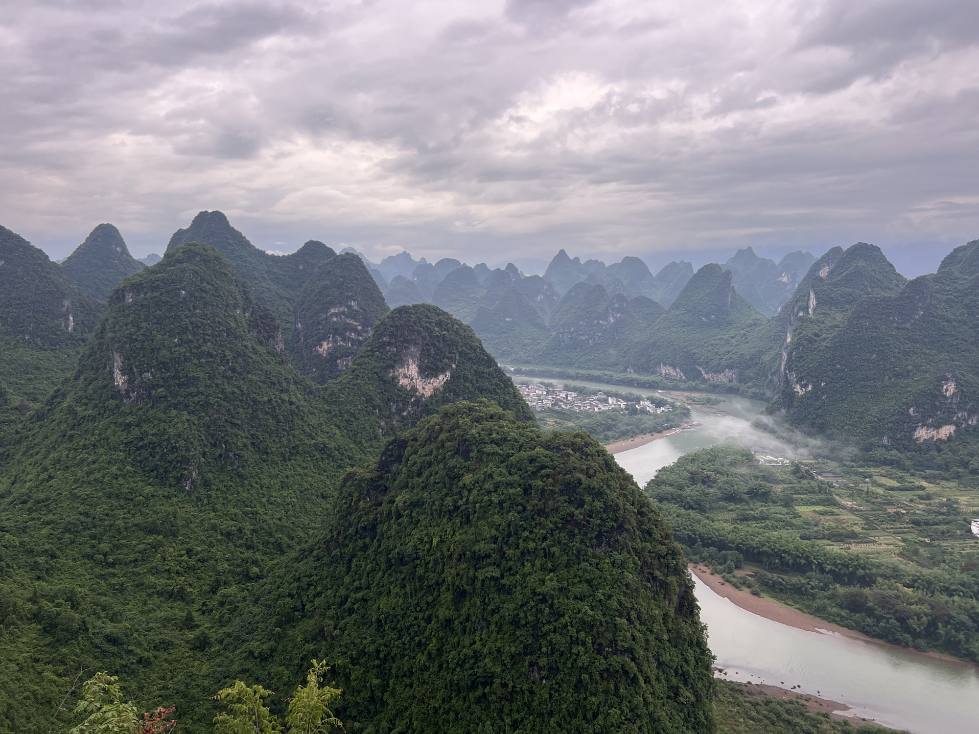 This landscape photograph depicts the karst mountains of Guilin, China under a cloudy sky. The foreground features lush green, cone-shaped hills, heavily vegetated with dense trees. A river meanders through the valley between the hills, with sandy banks visible in places. In the middle distance, there's a small village nestled among the hills. The background shows more distant karst formations, their outlines softened by atmospheric haze. The sky is overcast with layers of grey clouds. There are no people visible in the image. The general scene suggests a remote, natural landscape.