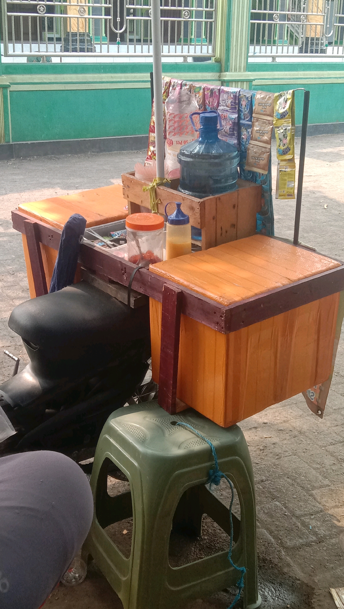 This is a medium shot image of a street food cart in Ngempit, Indonesia. The cart is built on a motorcycle chassis and features two wooden boxes with orange covers, one on each side. The left box is partially obscured by a motorcycle seat. On the right side of the cart, a large blue water jug with a blue cap sits on a wooden crate. Various packets of snacks or goods are hanging from a metal pole behind the water jug. A glass jar with a red lid, containing red spherical items, and a yellow bottle with a blue cap are placed on a wooden surface in front of the wooden crate. A dark blue cloth is draped over the left side of the cart. In the foreground, a green plastic stool is present, tied with a blue rope. A person's lower body, wearing a grey garment, is visible in the bottom left corner of the frame. The background shows a green wall with horizontal metal bars on top and a portion of a building. The ground is paved with dirt and loose stones.