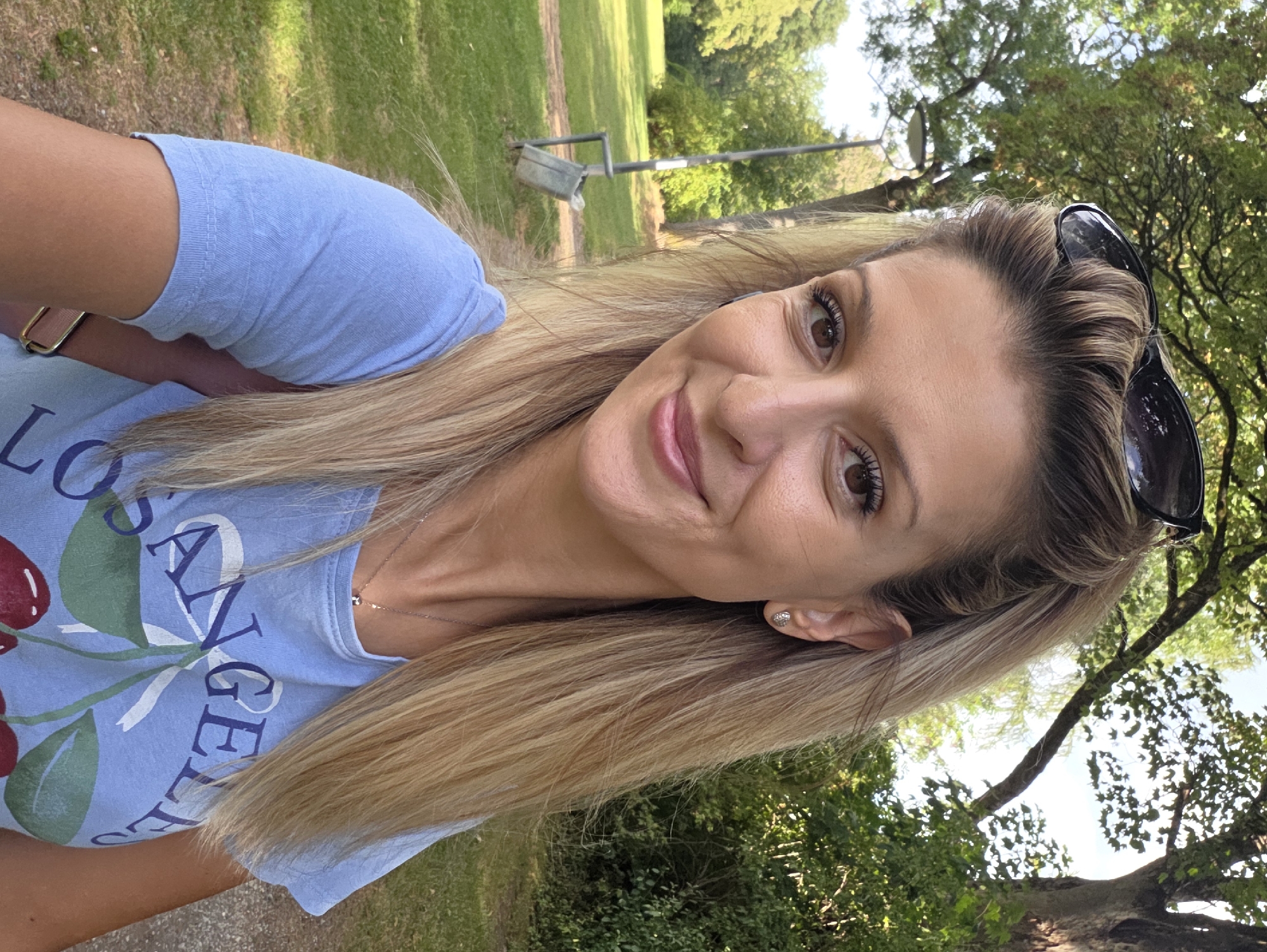 A woman is taking a selfie outdoors in a park. She has long blonde hair and is wearing a light blue t-shirt with a cherry graphic and the words "LOS ANGELES" printed on it. She is also wearing sunglasses on top of her head and a silver necklace. The background shows a grassy area, trees, and a lamppost with a trash can next to it. The lighting suggests it is daytime, likely in the afternoon, with sunlight filtering through the trees. The location is not identifiable as being in Munich, Germany.