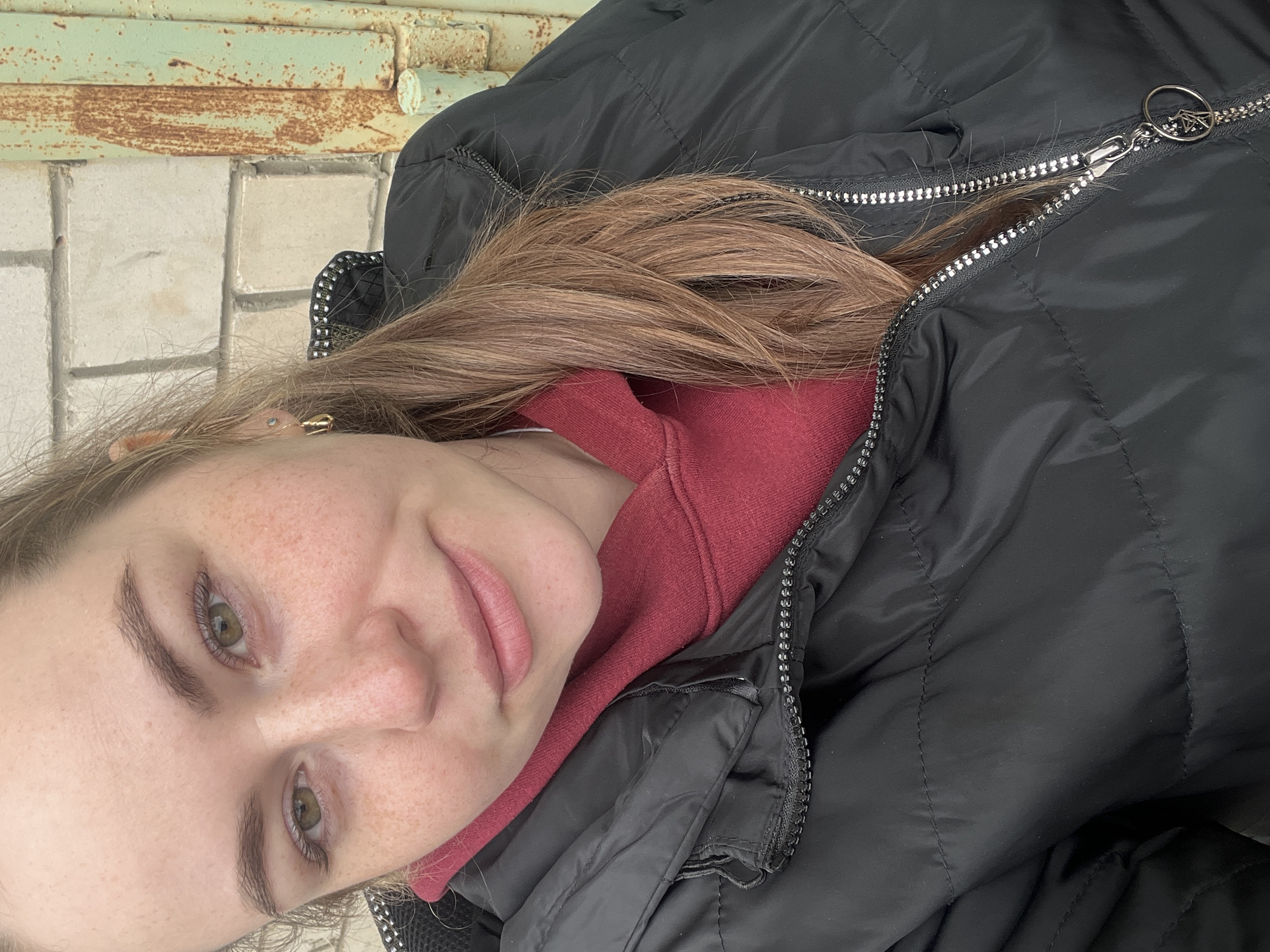 A young woman with fair skin, freckles, and light brown hair is wearing a black padded jacket and a red sweatshirt. She is outdoors, with a concrete sidewalk or patio and a rusty green metal structure in the background. It is likely she is in the city of Chaikovsky, Russia. The woman is looking directly at the camera with a slight smile. The photo is rotated counter-clockwise.
