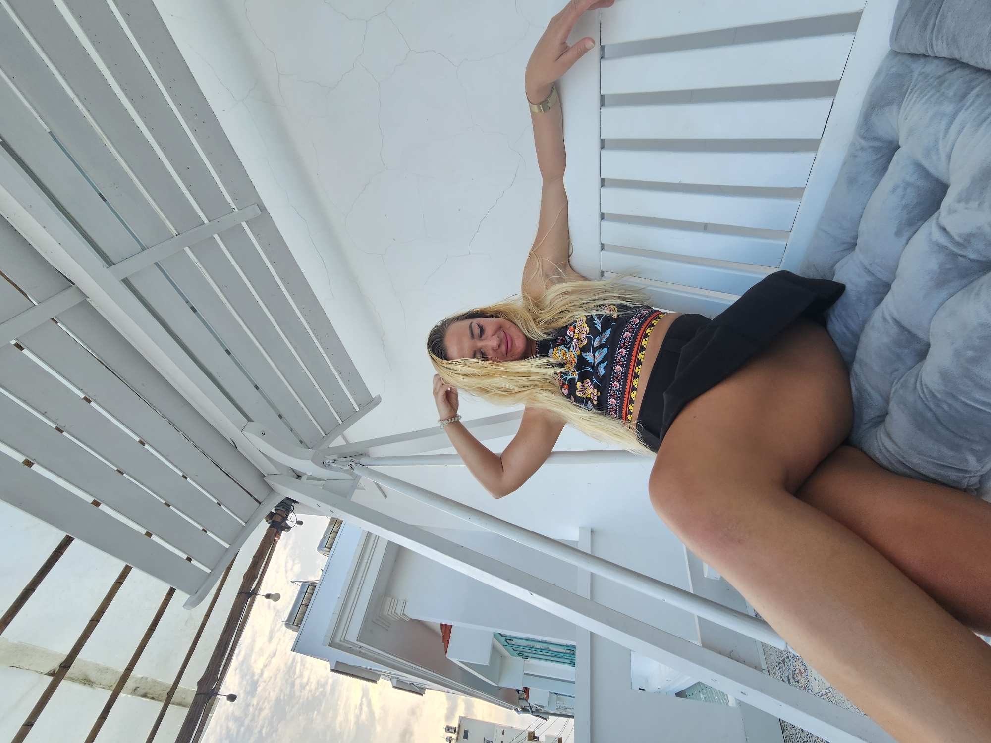 A woman with blonde hair and a patterned crop top and black skirt sits on a white swing with a tufted gray cushion. She is leaning back with one arm extended and her hand resting on the swing's backrest, while her other hand is raised and touching her head. The swing is positioned on a white tiled floor, with a white building and a cloudy sky visible in the background. The scene appears to be outdoors, possibly on a balcony or patio, in Phu Thuy Ward, Vietnam.