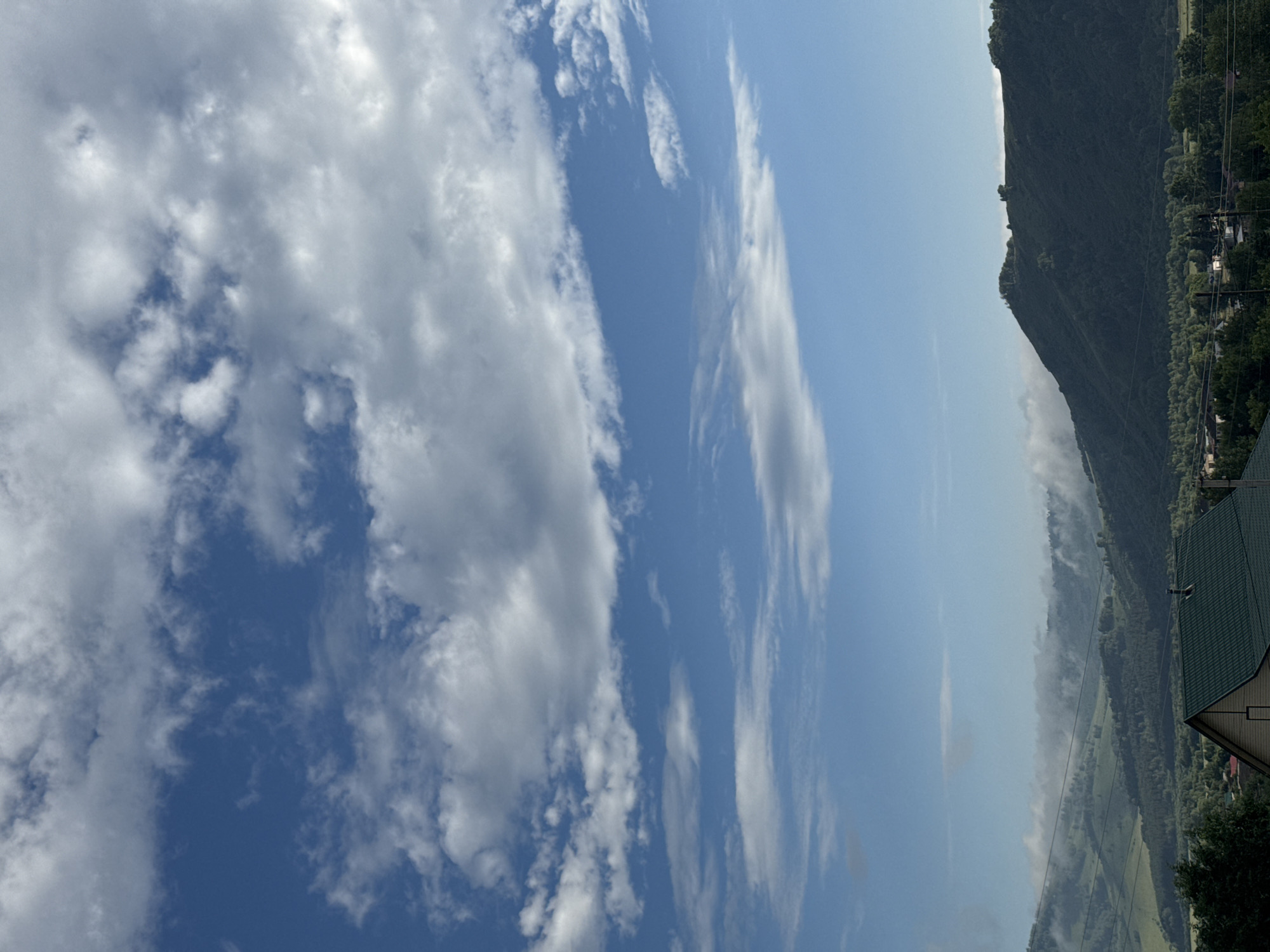 This is a vertical, wide-angle shot of a mountainous landscape in Soloneshnoye, Russia, on a bright, cloudy day. The sky occupies the majority of the frame, filled with a mix of fluffy white and wispy grey clouds against a vibrant blue backdrop. Sunlight illuminates the clouds, creating varying degrees of brightness and shadow.
To the right, a steep, densely forested mountain rises, its dark green slopes partially obscured by a hazy mist that clings to its base. Below the mountain, a valley unfolds, revealing patches of green fields and scattered trees. A few houses, some with dark green roofs, are visible nestled along the treeline and near the valley floor, hinting at a small settlement. Power lines crisscross the scene, adding a man-made element to the natural panorama.
The overall mood is serene and expansive, capturing the grandeur of the natural environment under a dynamic sky. The time of day appears to be midday, given the bright sunlight. There is no visible text in the image.