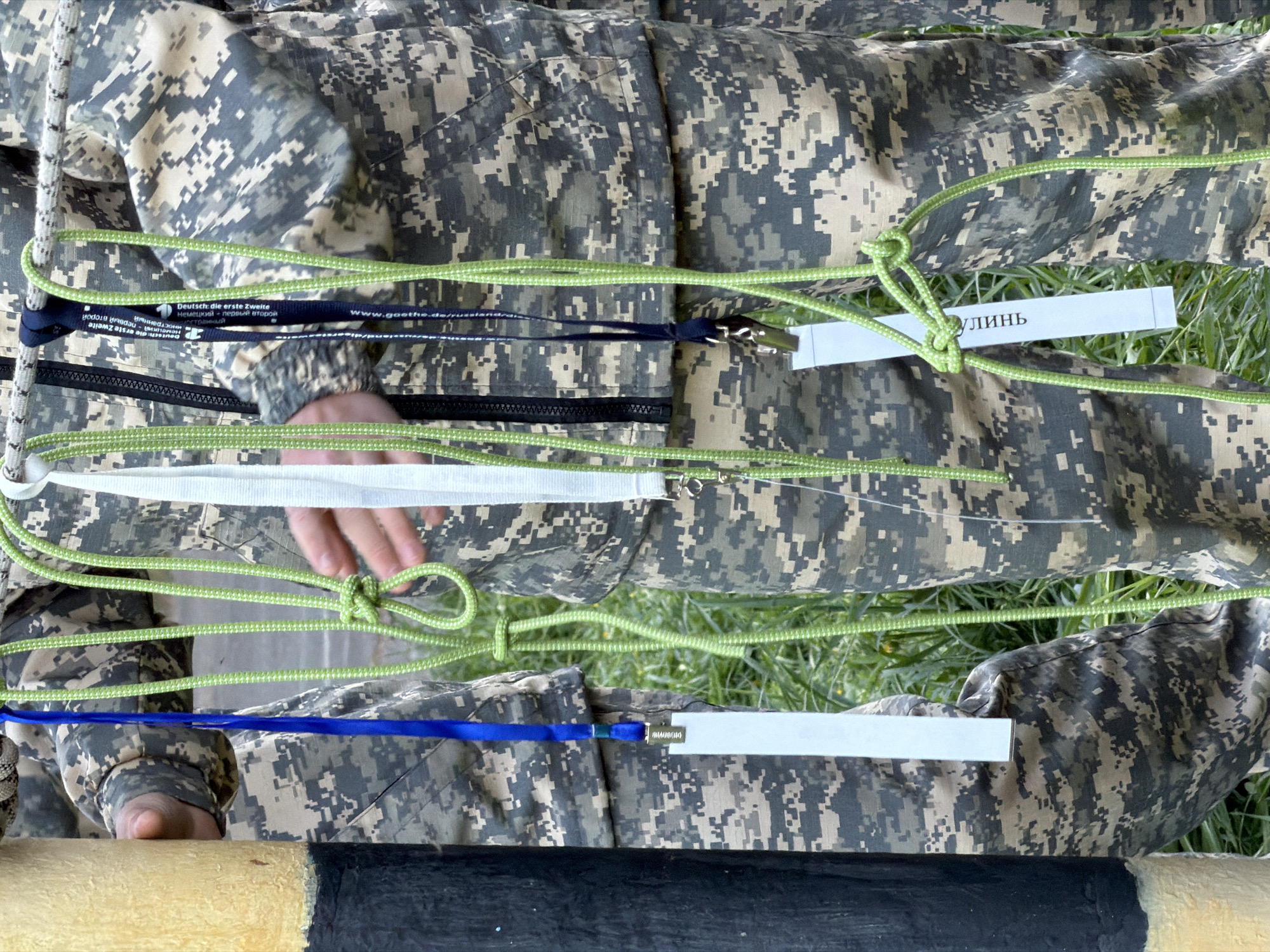The image depicts a person in camouflage clothing, likely a military uniform, seated or kneeling on grass. The person is interacting with multiple colored ropes, including light green, white, and blue. Some ropes are tied into knots. There are white rectangular tags attached to the ropes. One tag reads "ЛИНЬ" in Cyrillic script. Other text is visible on one of the ropes which appears to be "Deutsch: die erste Zweite", followed by "Немецкий первый второй". The bottom of the image includes a striped beam painted with cream and black. The setting is likely outdoors in Забегалово, Russia.