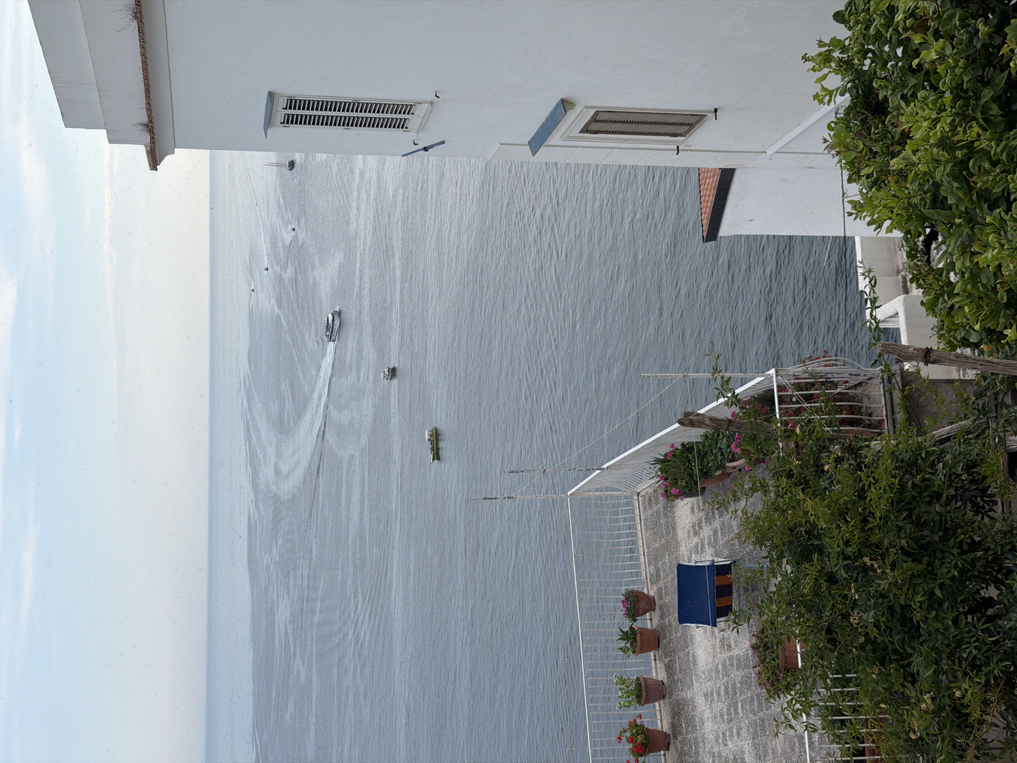 From a vantage point in Pogerola, Italy, the image captures a vista overlooking the ocean. The foreground features a terrace with potted plants and a blue and brown striped chair. A white railing borders the terrace, and foliage from surrounding trees and shrubs intrudes into the frame on the right. The building's white exterior, complete with ventilation grilles, is visible above the terrace. In the distance, a vast expanse of ocean stretches out, dotted with several small boats. One boat is moving, leaving a distinct wake in the water. The sky is pale blue, indicating a clear day.