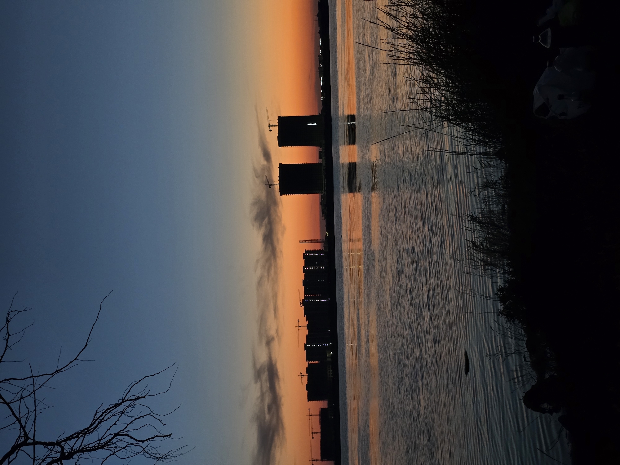 The image captures a sunset over the city of Yevpatoriya, Ukraine. In the foreground, dark silhouettes of grass and brush line the edge of a body of water. The water is calm, reflecting the vibrant hues of the sunset. In the mid-ground, a series of buildings are silhouetted against the fiery orange and yellow sky. Some of these structures appear to be under construction, with cranes visible. Two tall, cylindrical buildings stand out, one of which displays a vertical line of illuminated dots. The sky transitions from a deep blue at the top to bright orange and yellow near the horizon. A bare tree branch extends from the upper left corner of the frame, silhouetted against the sky. The overall scene conveys a sense of twilight tranquility amidst ongoing urban development.