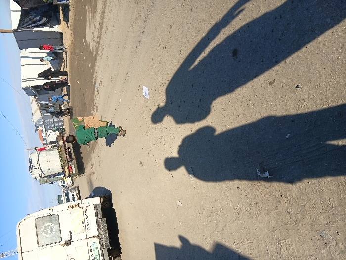 A boy in a green outfit carries boxes on a street lined with white tents and parked trucks. Several people stand near the tents. The shadows of two people are cast onto the road, suggesting the presence of someone taking the photograph. The road appears dry and dusty with some visible debris. The sky is clear blue.