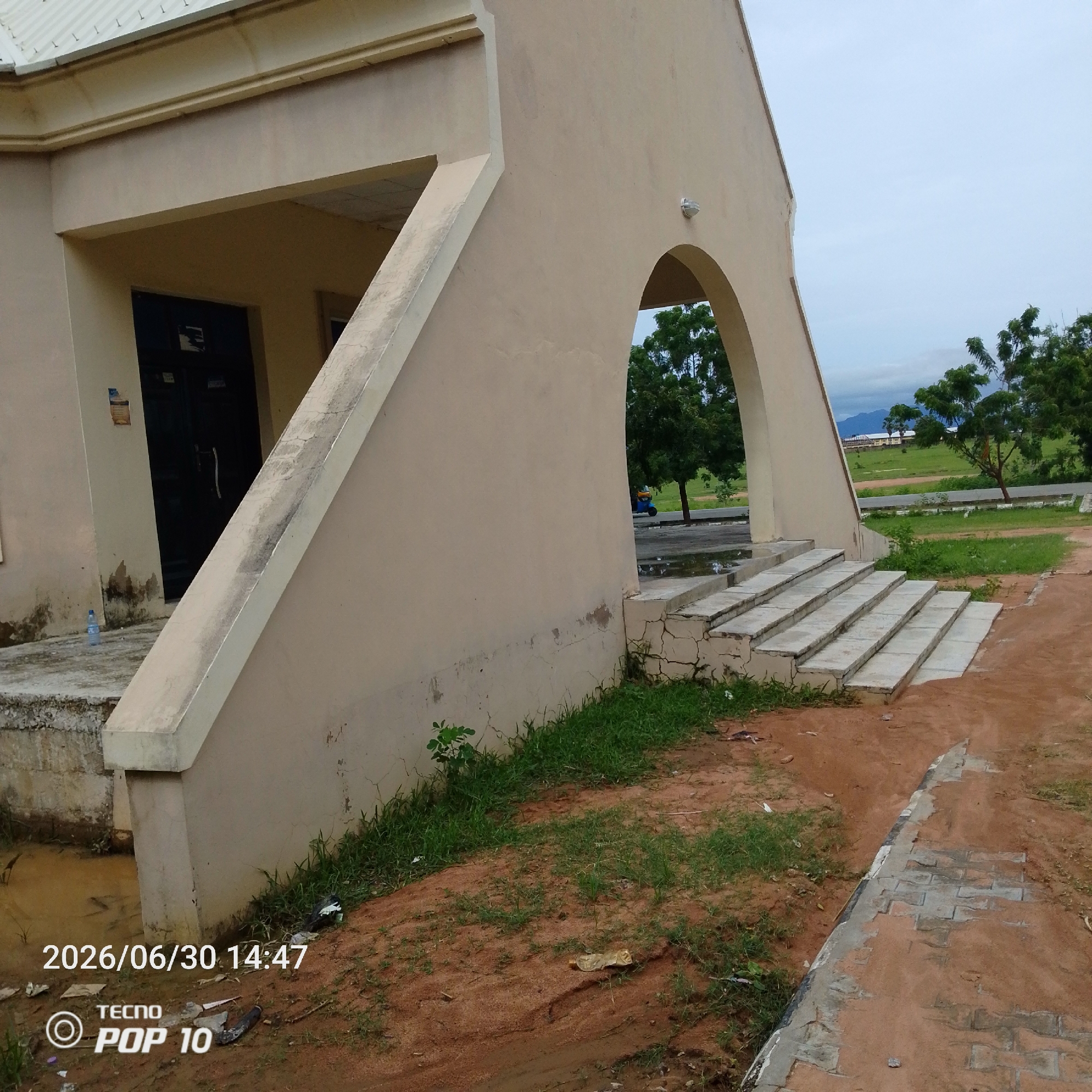 The image shows the exterior of a light-tan, modern building with an arched entryway and a slanted support structure. In front of the building, there are a set of concrete steps leading up to the arch. The ground is a mix of sandy soil, sparse green grass, and some areas with standing water, suggesting it may have recently rained. To the left of the steps, a plastic water bottle is visible on a concrete slab. Beyond the arched entryway, trees and a green landscape are visible, with hills or mountains in the far distance under a cloudy sky. The scene appears to be an outdoor area on a slightly overcast day.
Visible text includes a timestamp "2026/06/30 14:47" and a logo with "TECNO POP 10". There are also some smudges and markings on the building's walls and the surrounding ground. The overall impression is of a building in a semi-urban or rural setting in Jalingo, Nigeria, possibly a public space or an educational institution. No people are visible in the image, and the focus is on the architecture and the immediate surroundings.