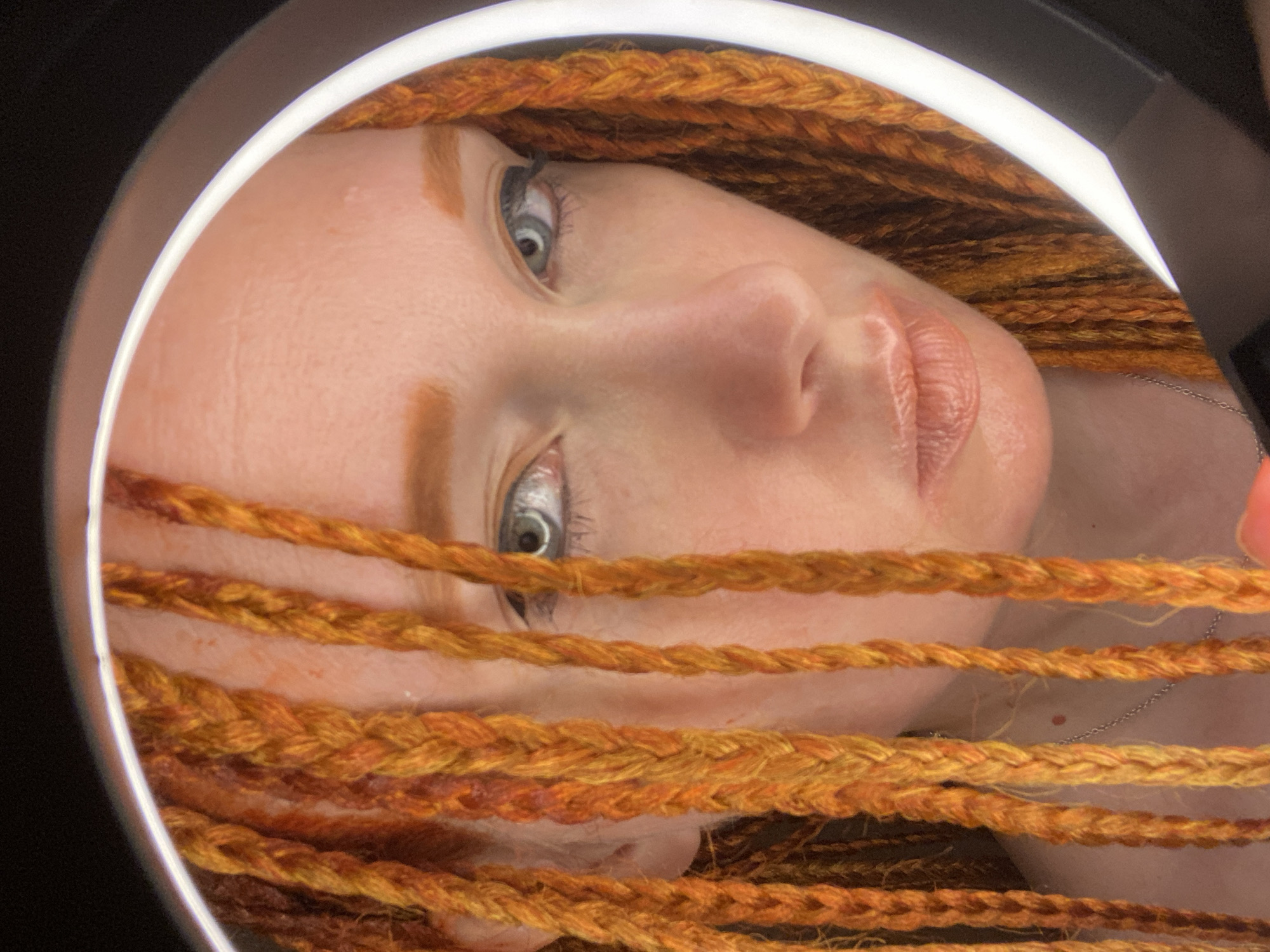 This is a close-up, self-portrait taken indoors. A person with orange braided hair is looking towards the camera. Their face is partially framed by a circular light, suggesting it might be a ring light used for photography or video. The background is dark and out of focus. The person's eyes are open, and they are wearing light makeup, including eyeliner. There is a faint necklace visible on their neck. The lighting creates a soft glow on their skin. The image does not contain any discernible information related to the city of Sochi, Russia.